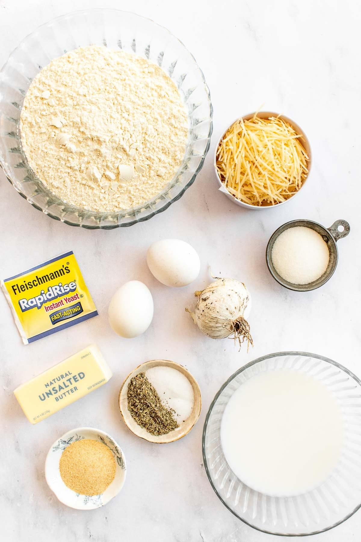 Bowls of cheese, flour, milk, spices, and sugar next to eggs and butter on a table.