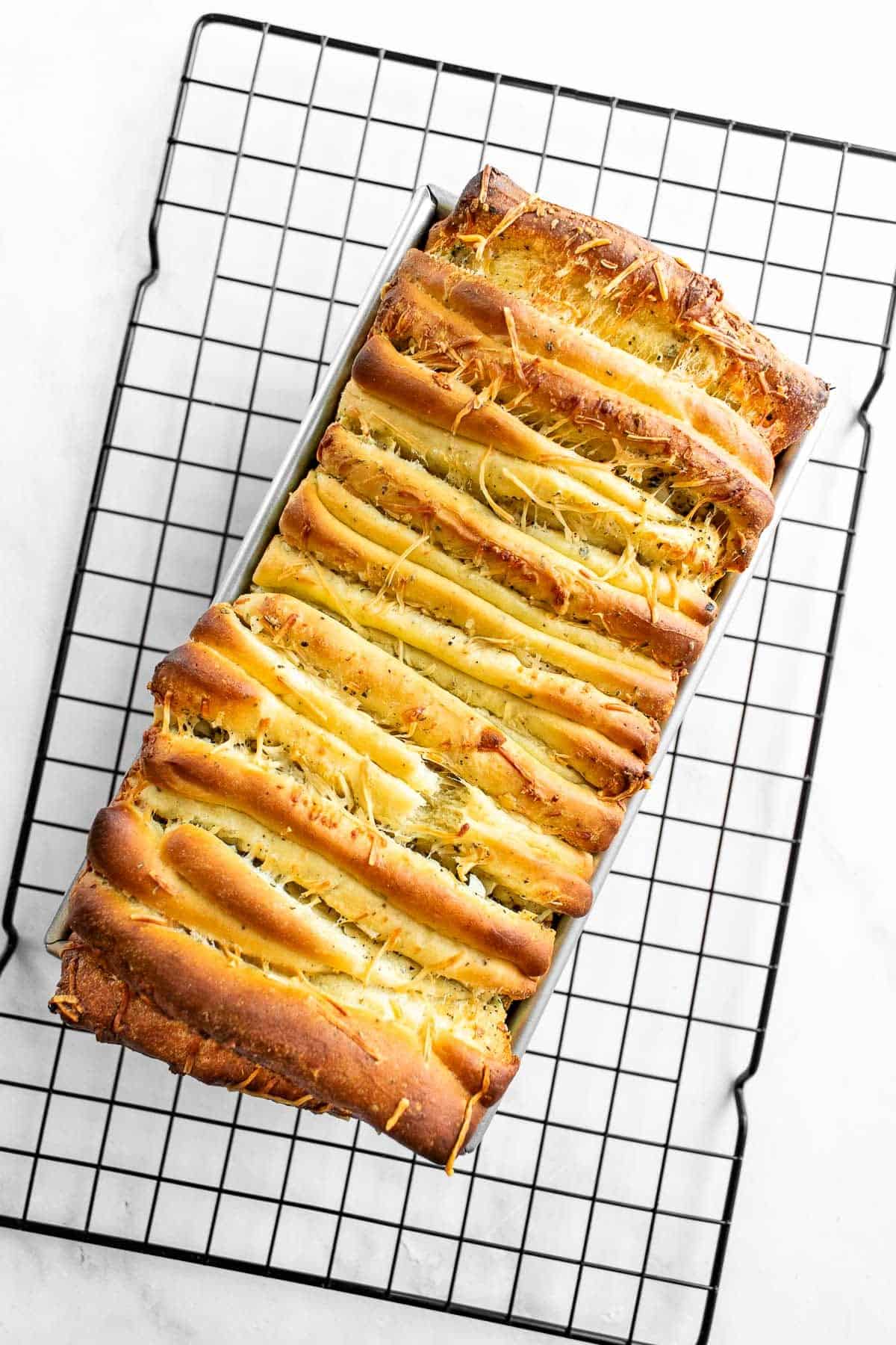 A baked loaf of garlic pull apart bread in a loaf pan on a cooling rack.