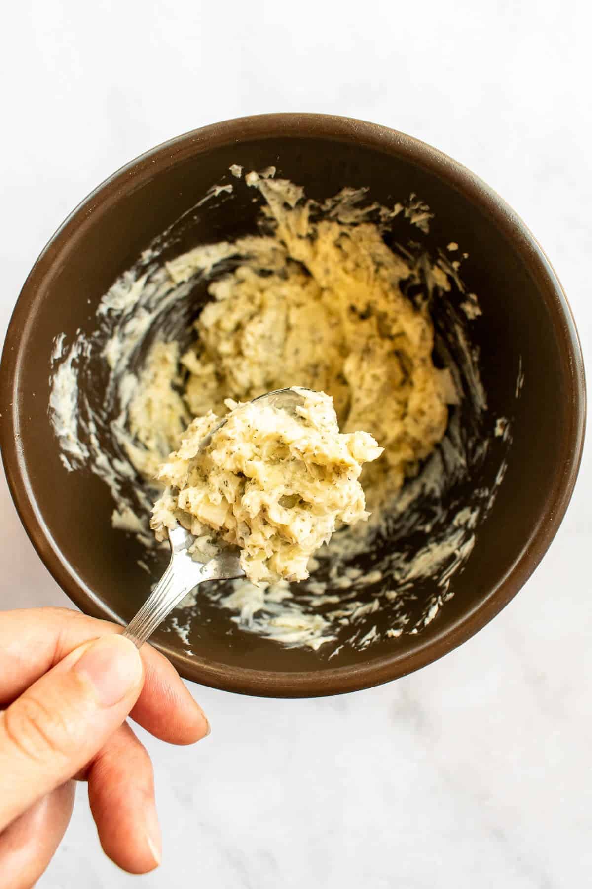 A hand mixing butter with garlic powder and dried herbs in a bowl.