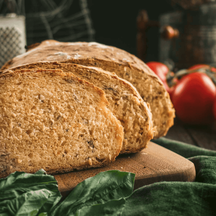 Two slices of tomato bread with flecks of dried basil inside leaning against the rest of the sliced loaf on a wood board.