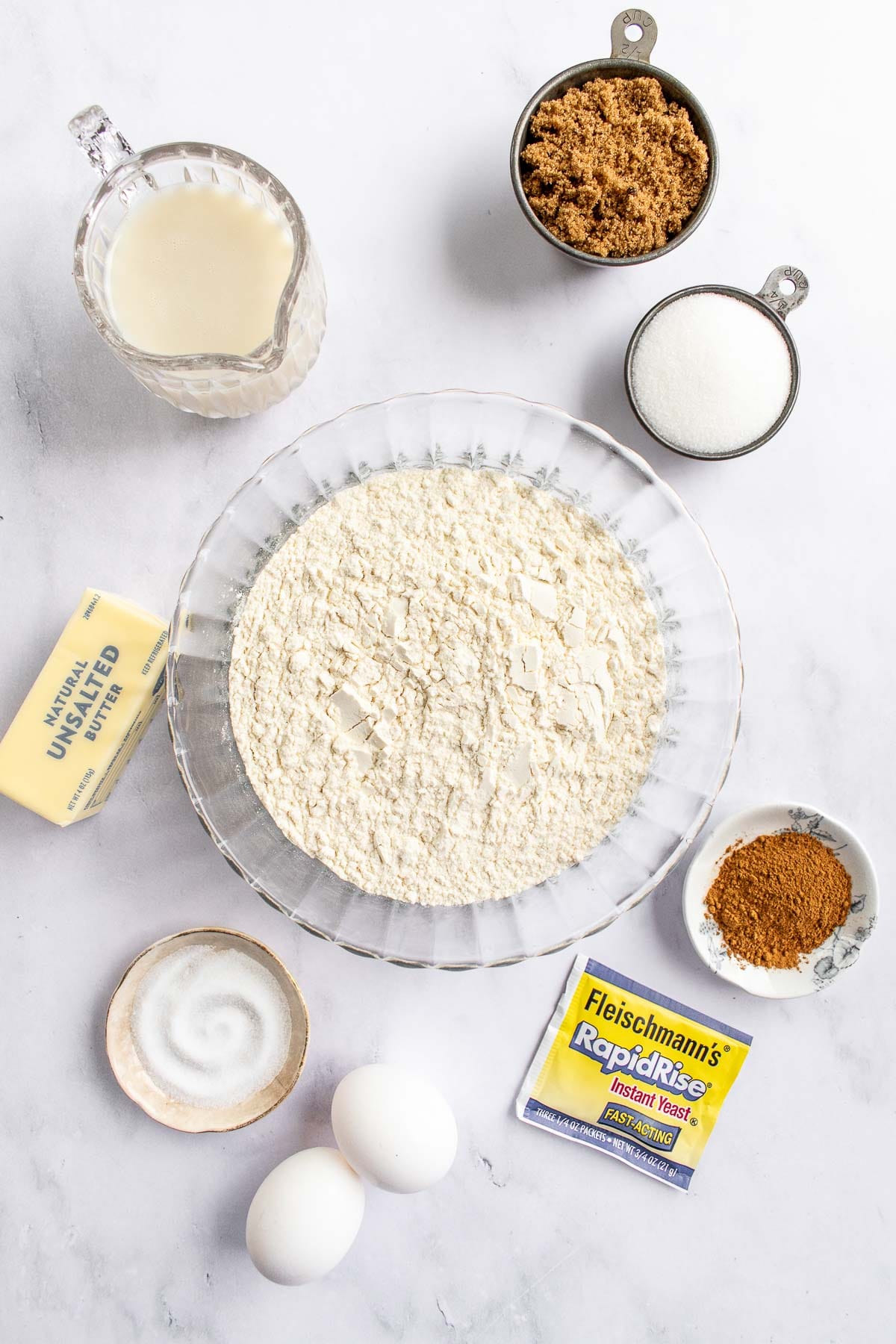 Bowls of flour, cinnamon, sugar, salt, and milk on a table next to butter and yeast.