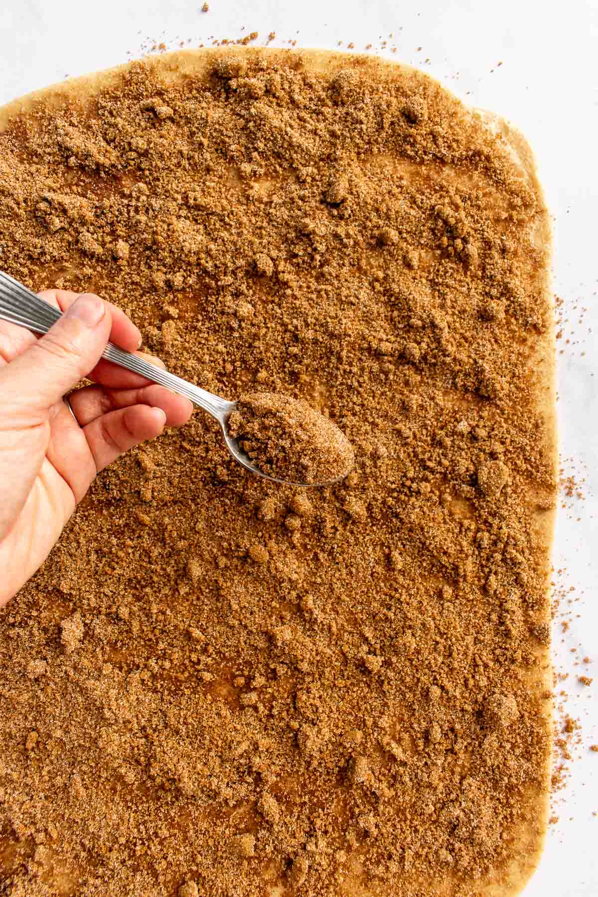 A hand sprinkling cinnamon sugar onto dough with a spoon.