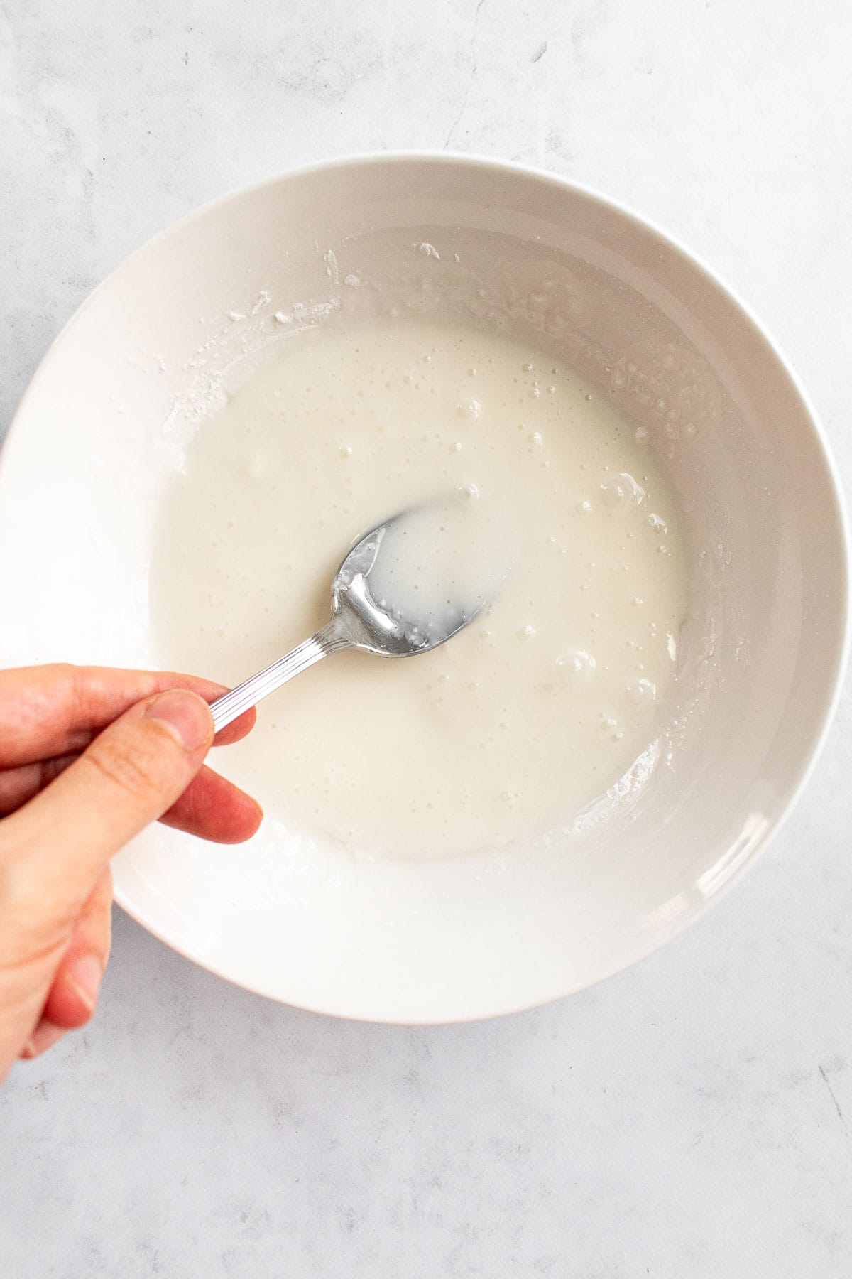 A hand mixing white glaze in a bowl with a spoon.