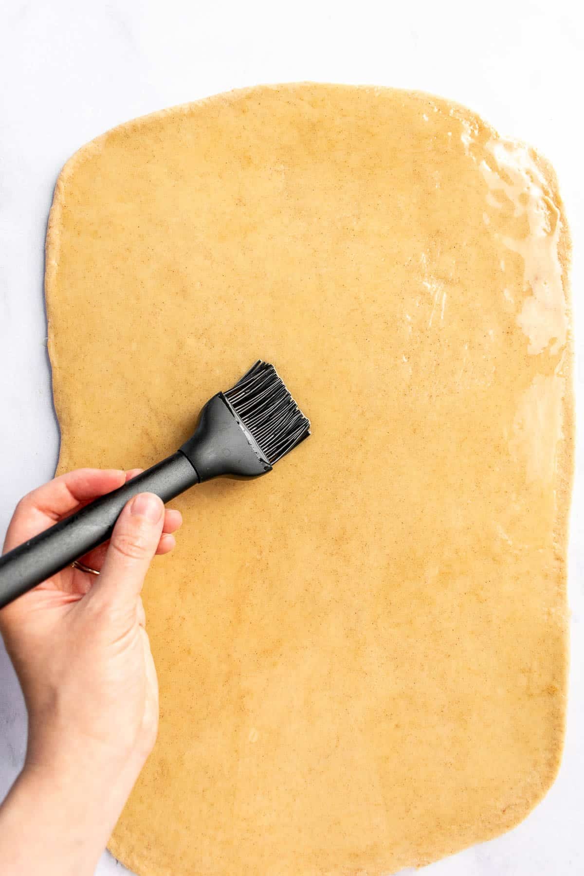 A hand brushing melted butter on rolled-out bread dough.