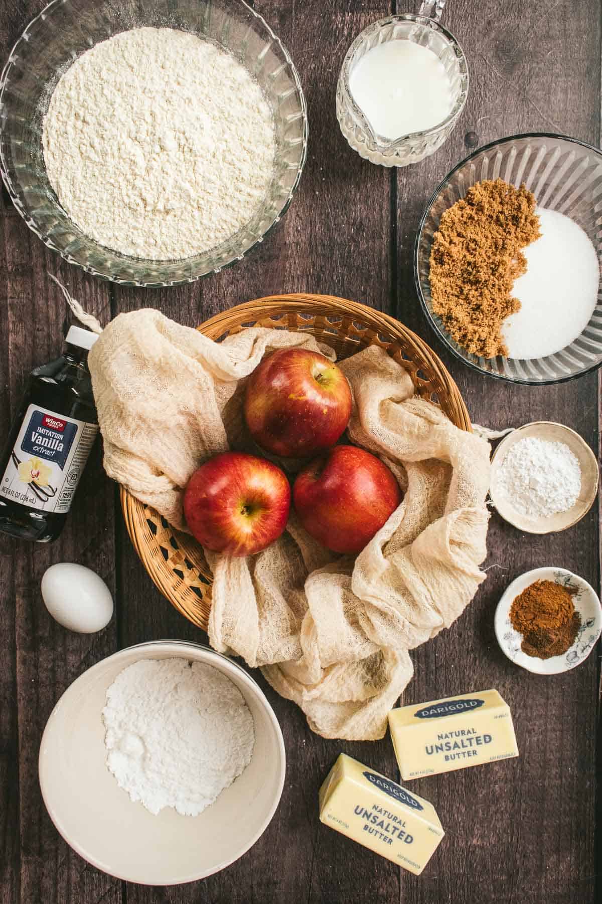 A basket of apples next to bowls of flour, sugar, spices, salt, and sticks of butter.