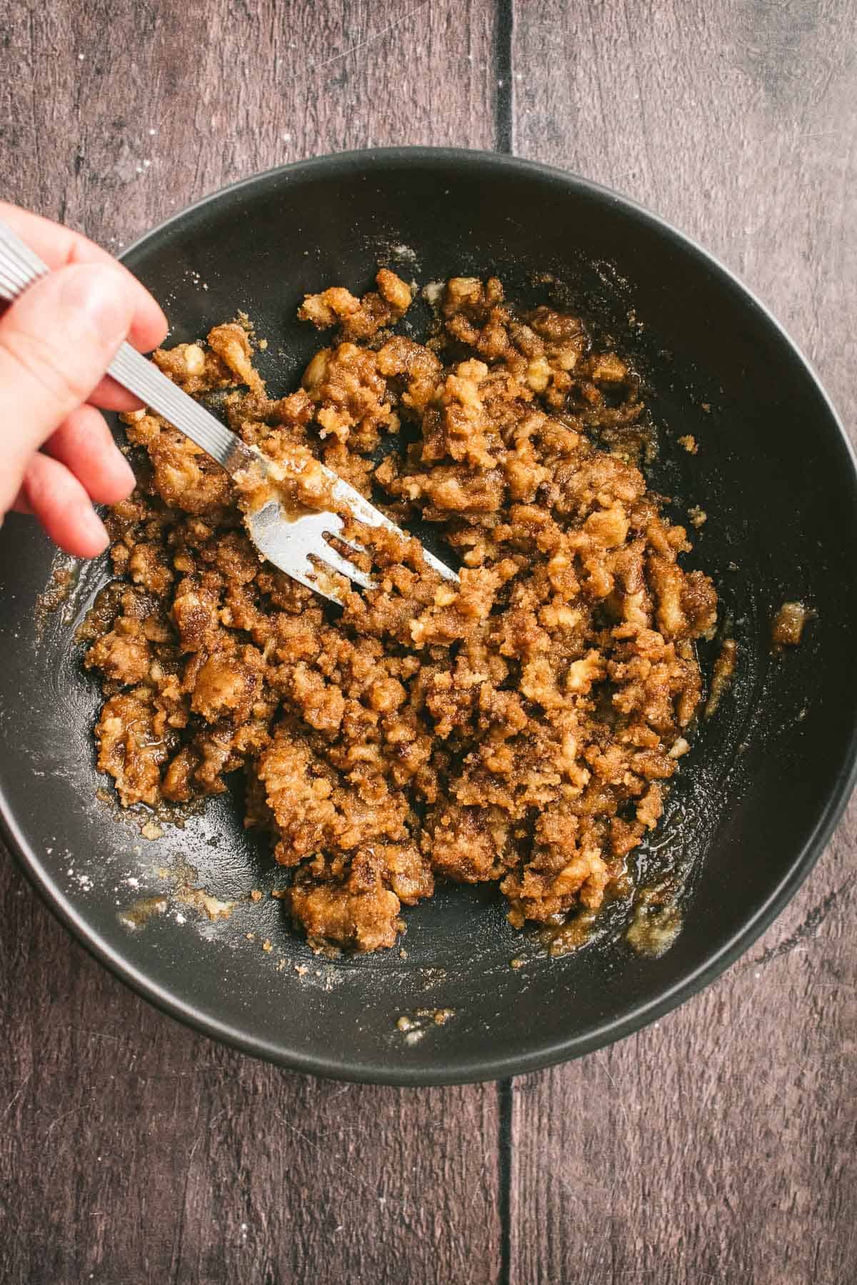 A hand mixing a crumbly brown cake topping in a black bowl with a fork.