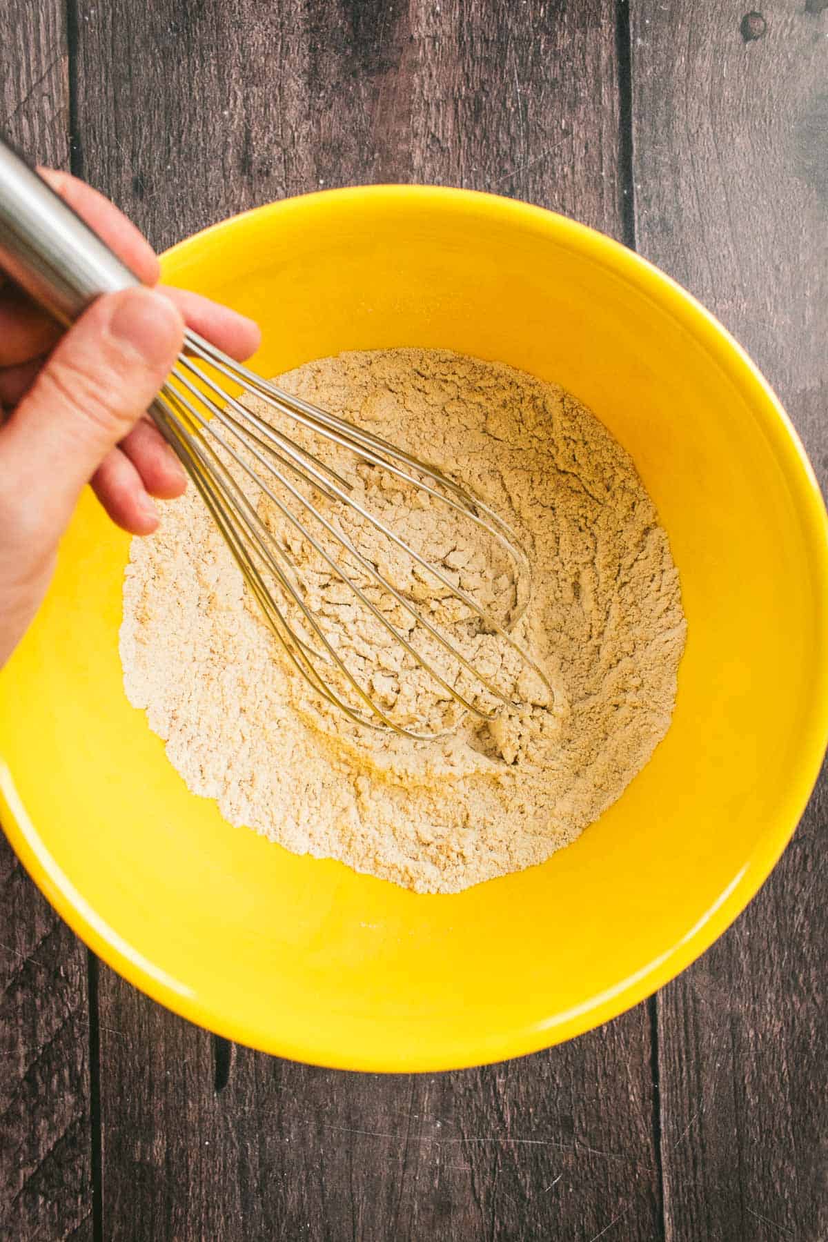 A hand whisking flour and baking spices in a yellow bowl.