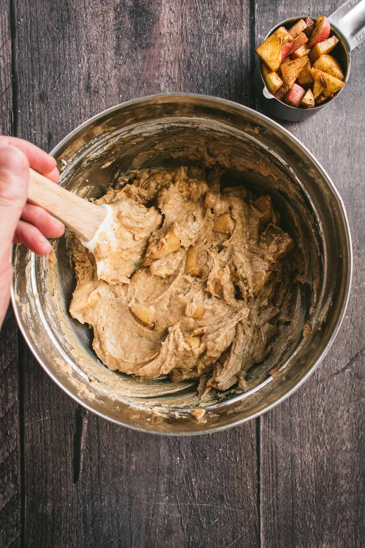 A hand stirring apple cake batter in a mixing bowl.