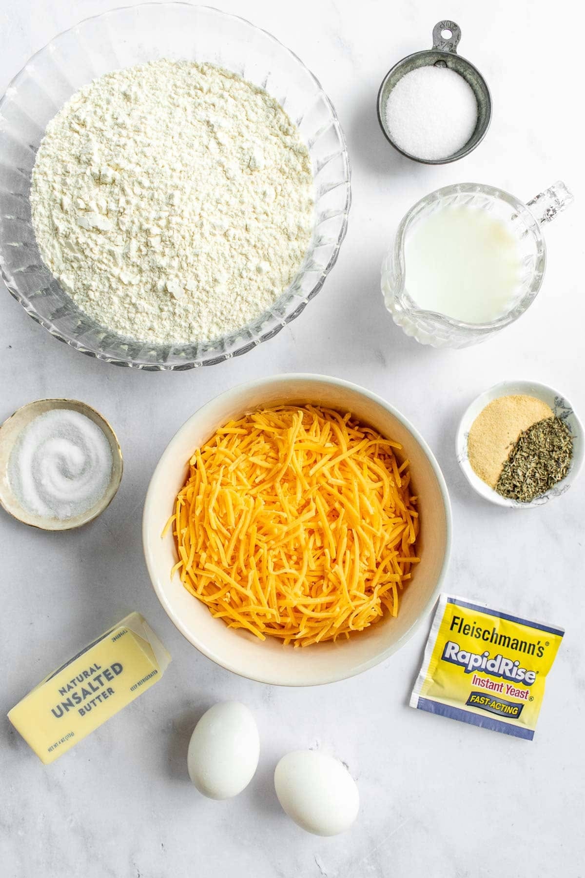Bowls of flour, milk, sugar, spices, and cheddar cheese next to eggs, butter, and a packet of yeast. 