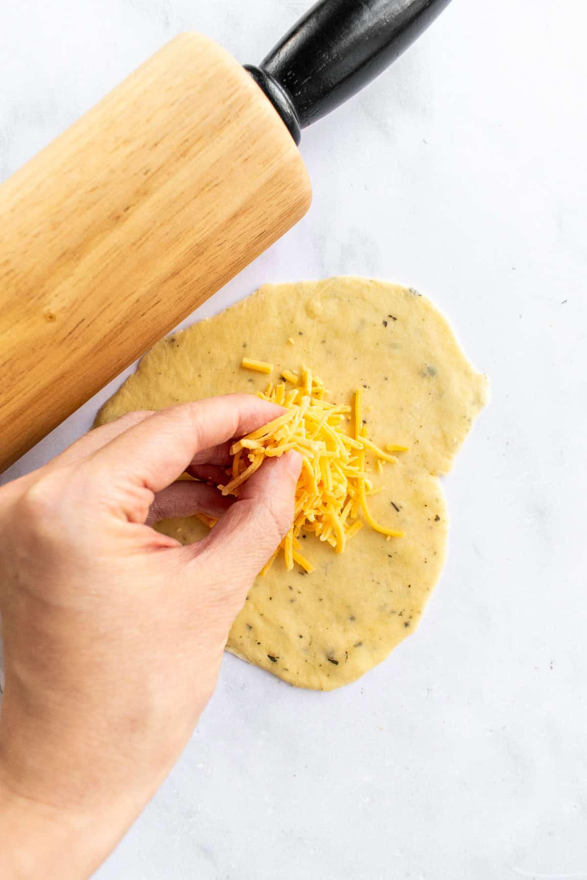 A hand sprinkling shredded cheese on a circle of bread dough. 