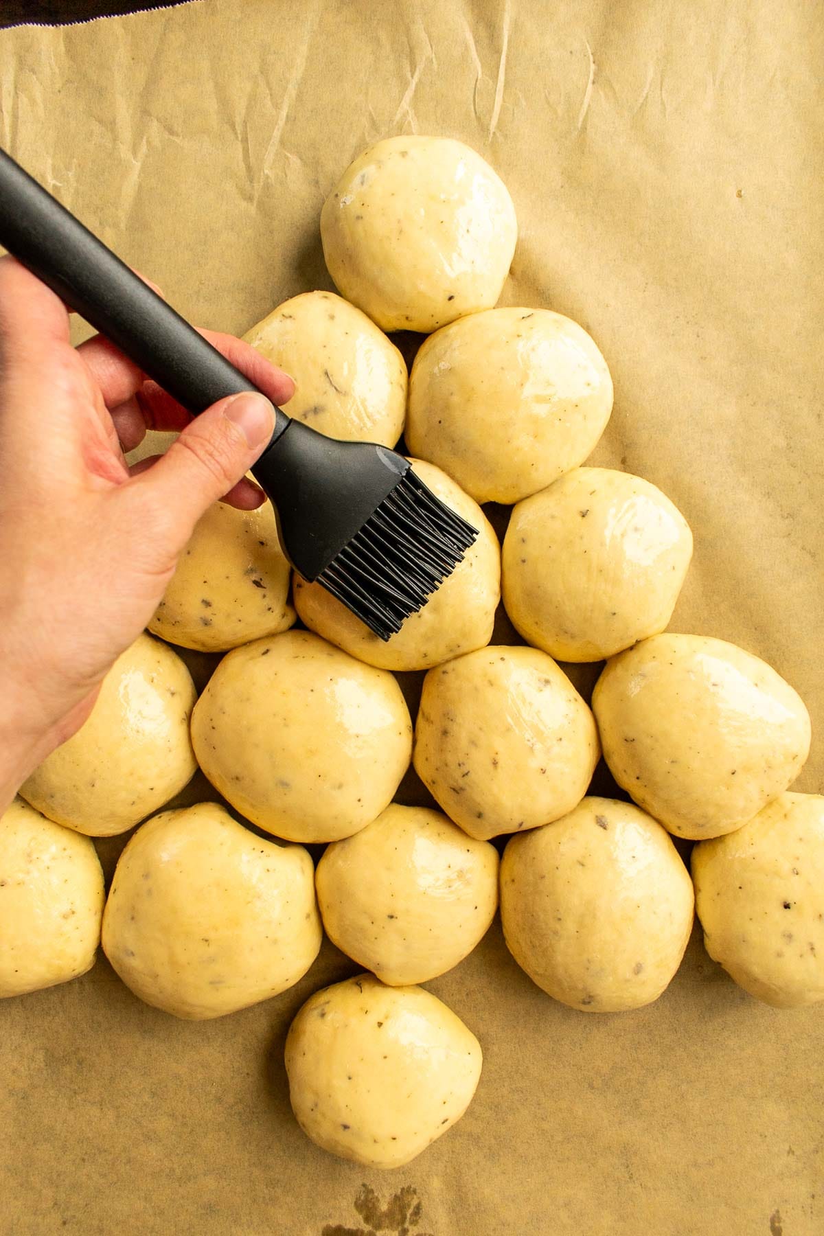 Bread buns on a baking sheet being brushed with an egg wash. 