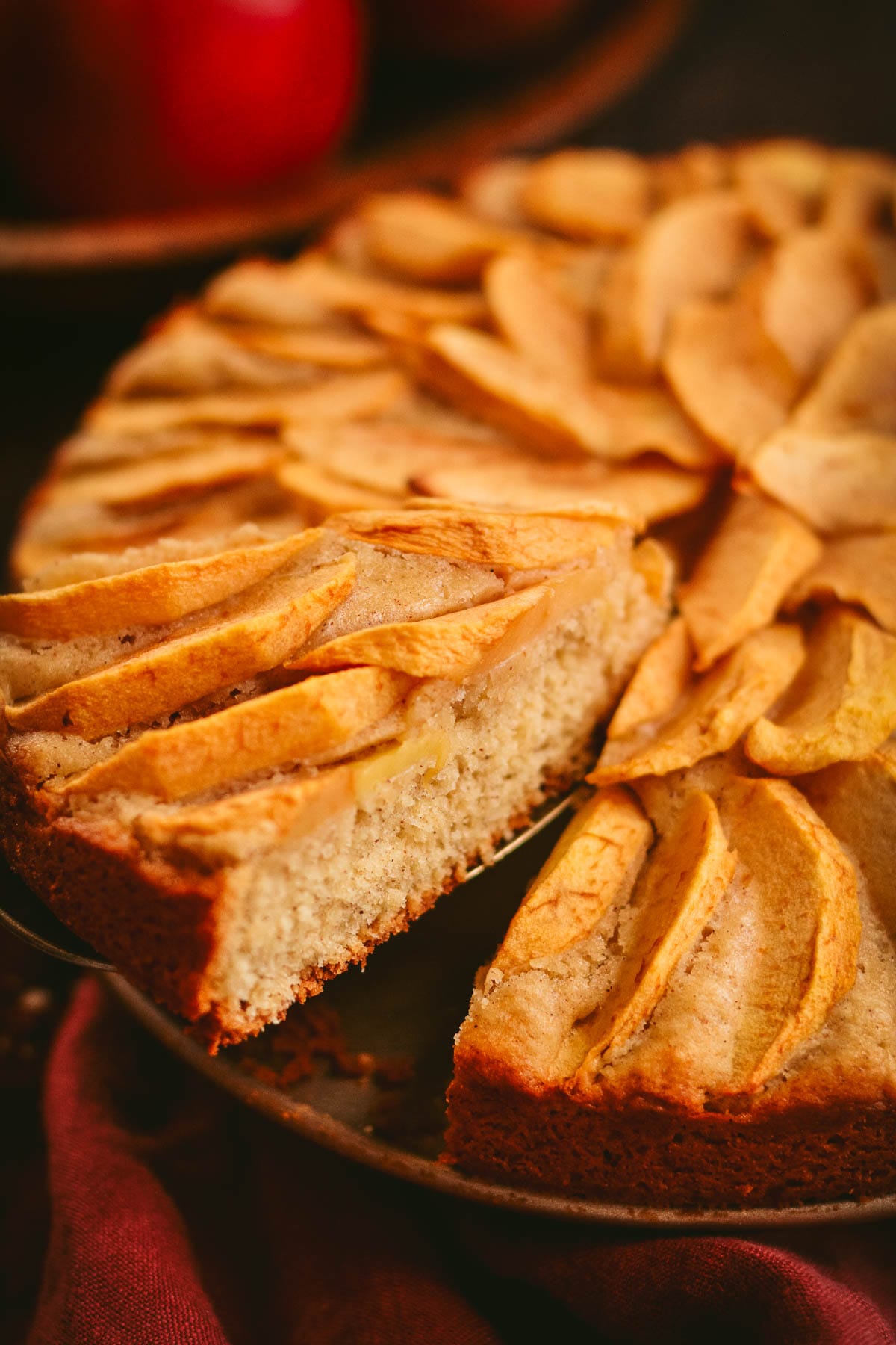 A buttery cake topped with peeled sliced apples on a red linen.