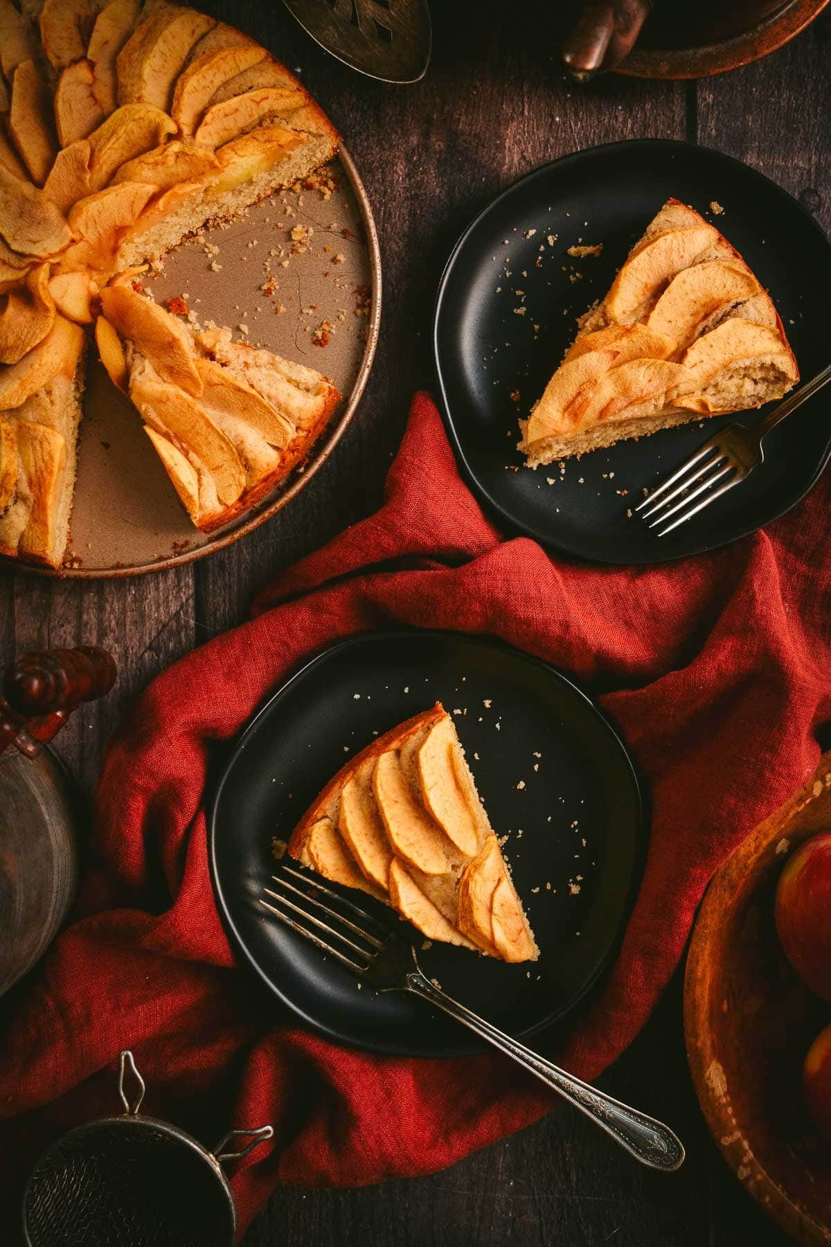 Two slices of apple cake on plates next to the rest of the cake.