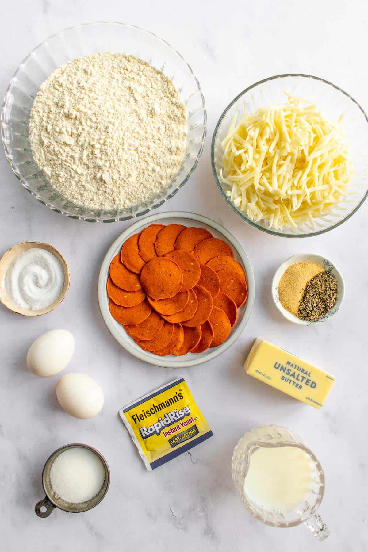 Bowls of flour, yeast, cheese, pepperoni, sugar, salt, and milk next to eggs and a stick of butter.
