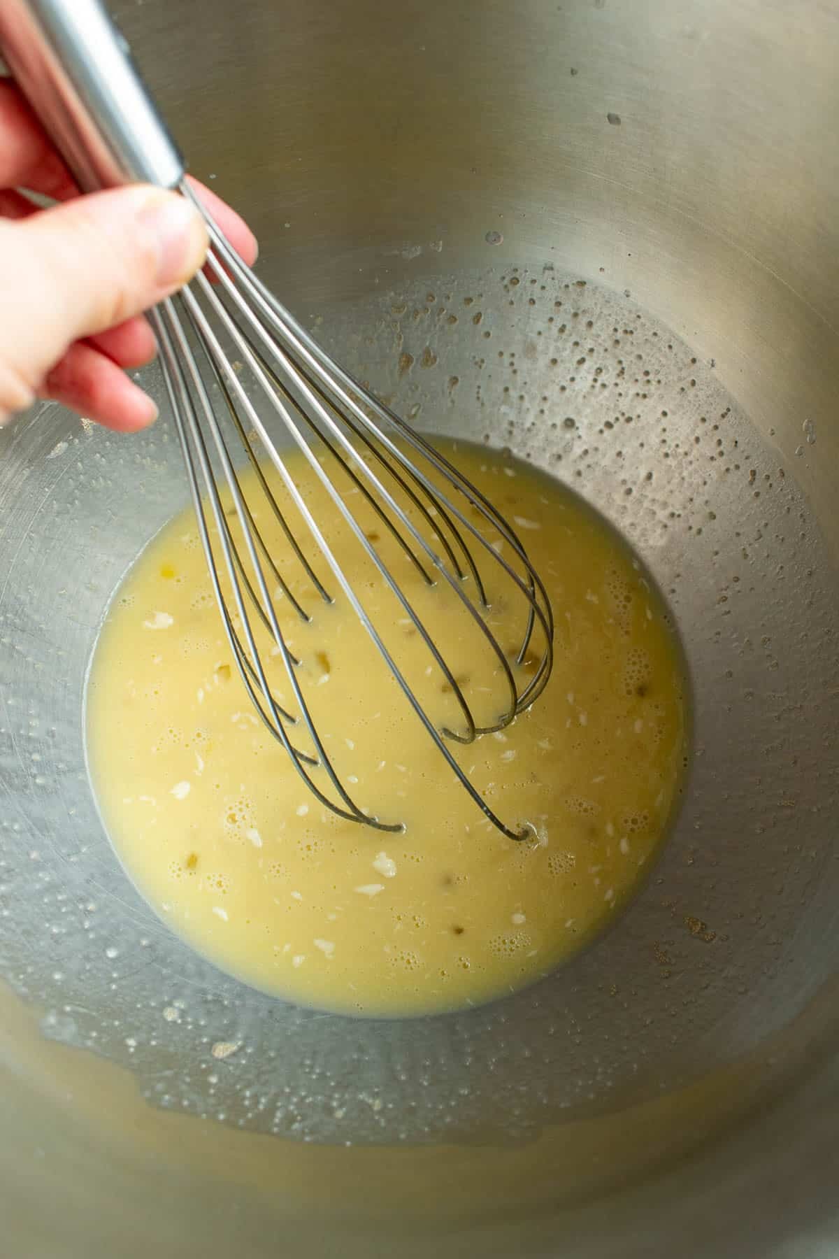 A hand whisking milk, sugar, yeast, and butter in a metal bowl.
