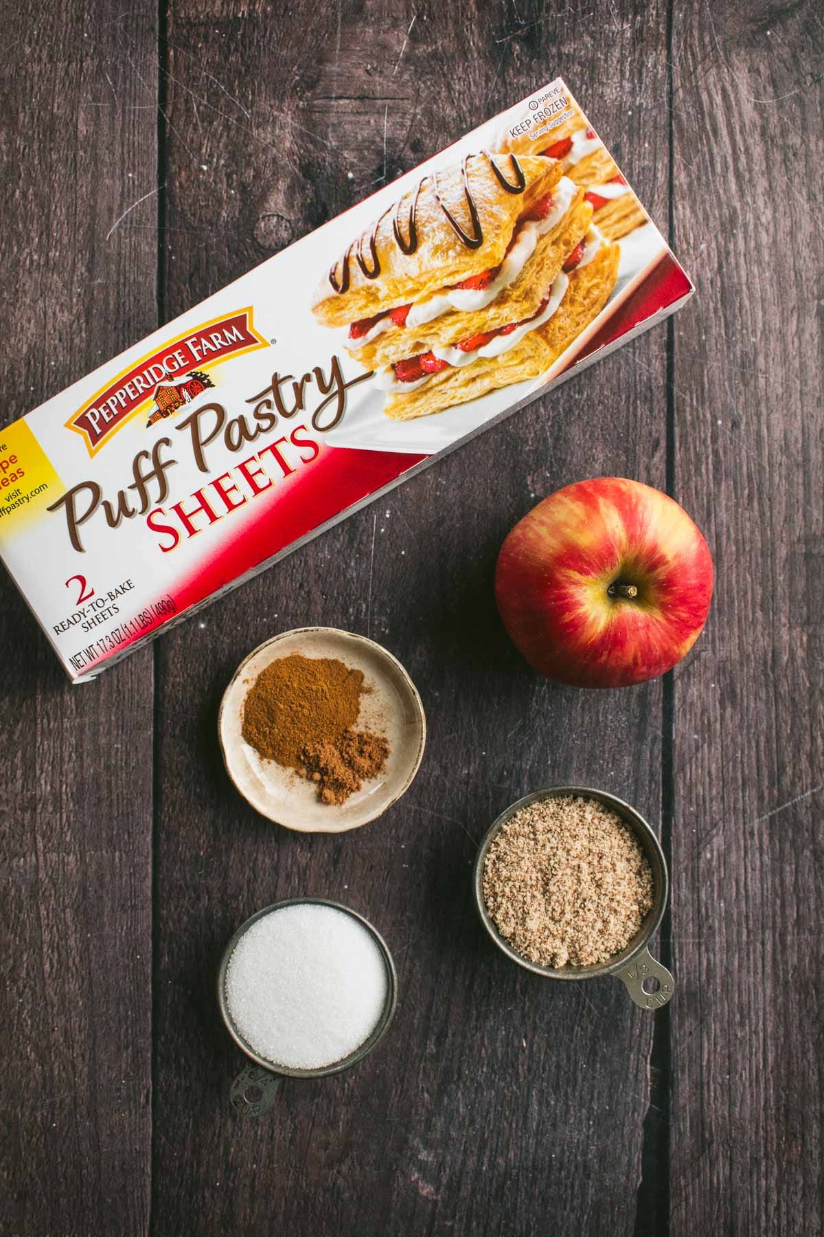 A box of puff pastry and an apple on a table next to bowls of sugar, cinnamon, and almond flour.