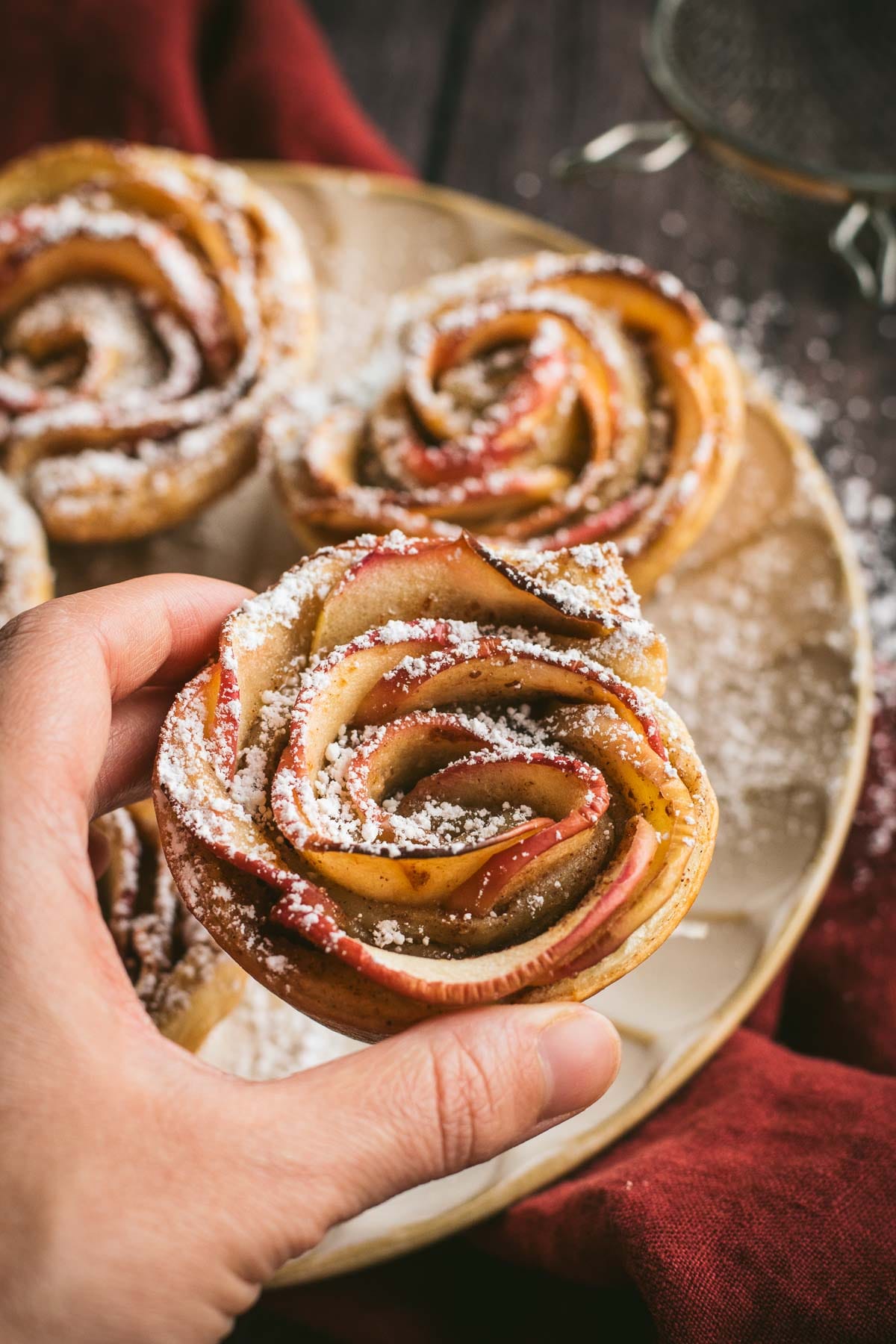 A hand holding an apple rose pastry dusted with powdered sugar.