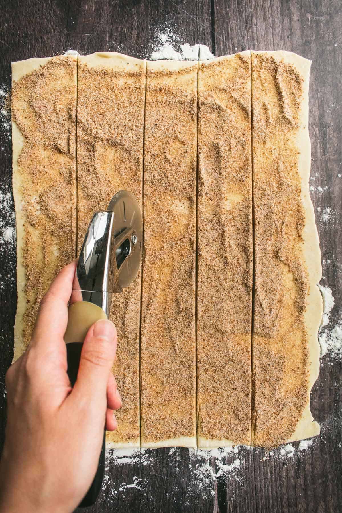 A hand slicing puff pastry into strips with a pizza cutter.
