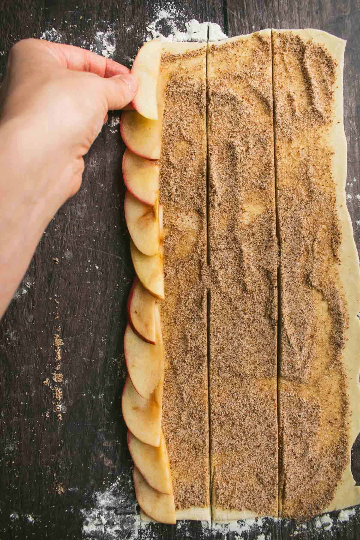 A hand layering apple slices on a strip of pastry dough. 