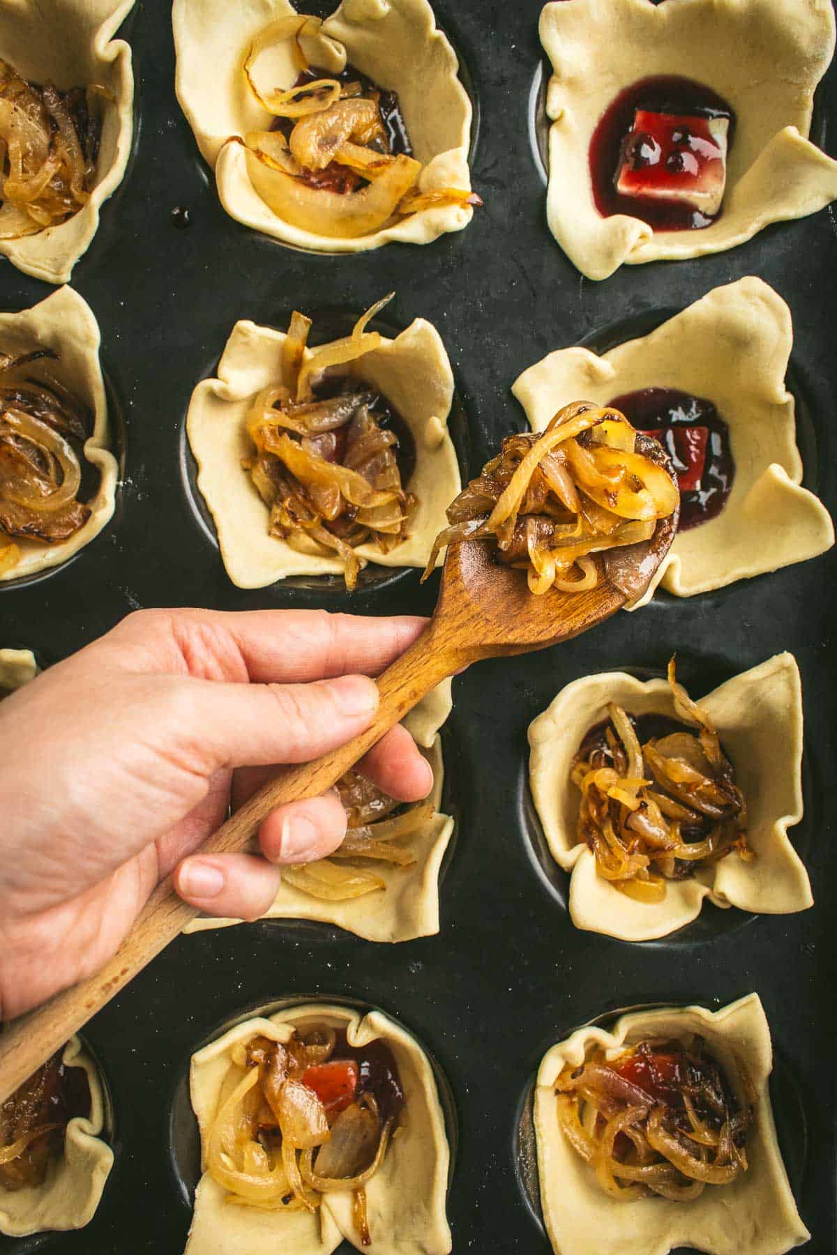 A hand spooning caramelized jam onto squares of pastry dough in a muffin tin. 