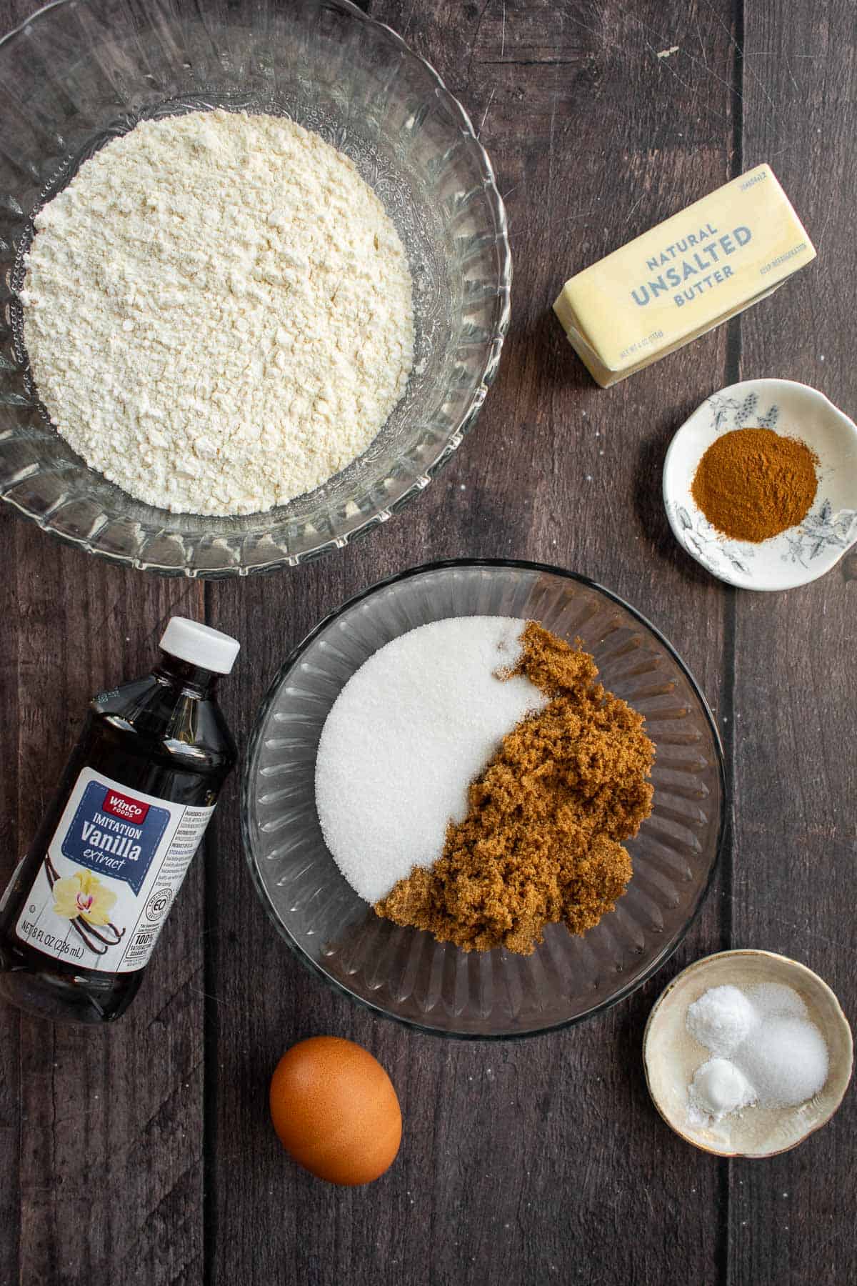 Bowls of flour, sugar, and spices next to an egg, a stick of butter, and a bottle of vanilla.