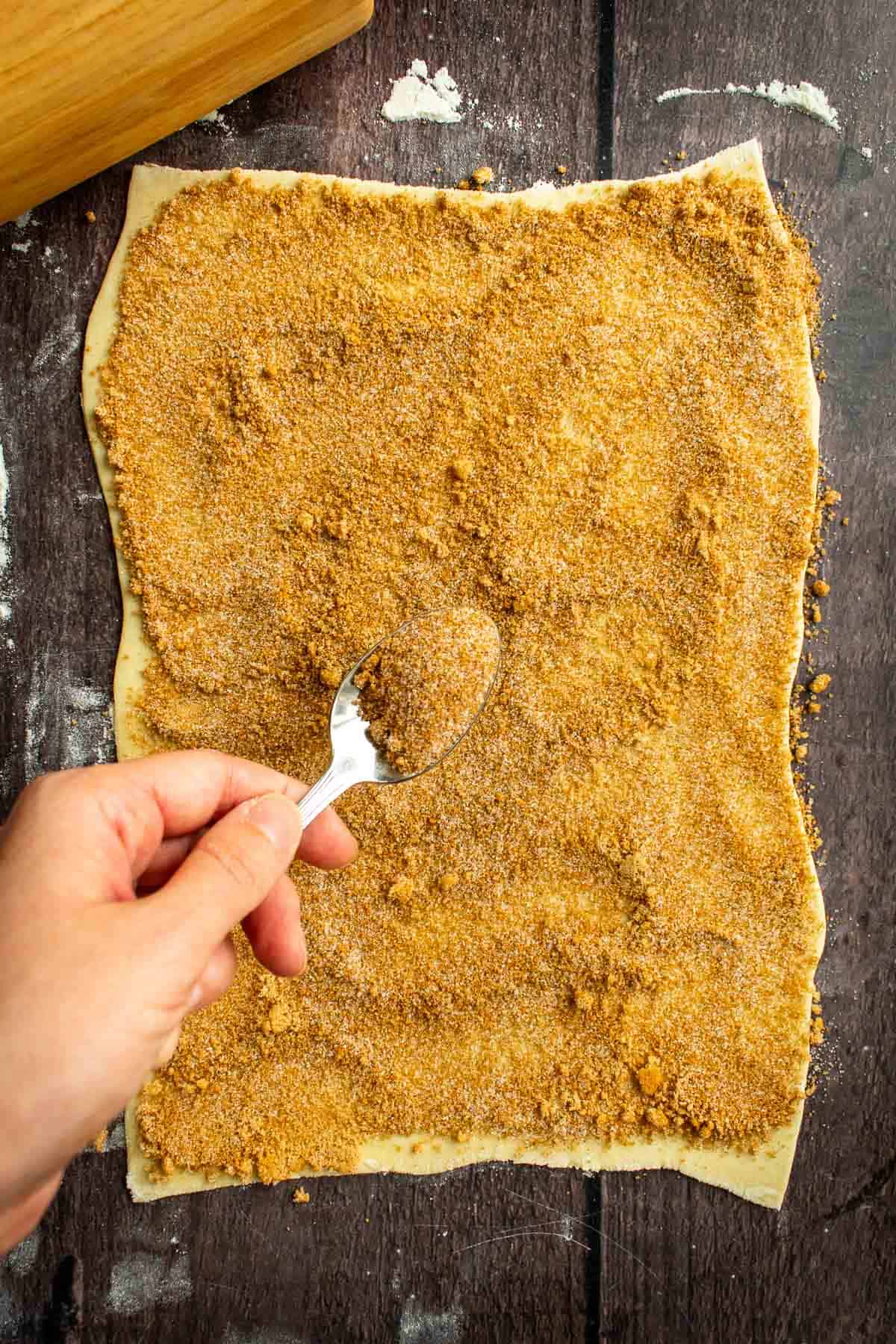 A hand spreading cinnamon sugar over rolled-out puff pastry. 