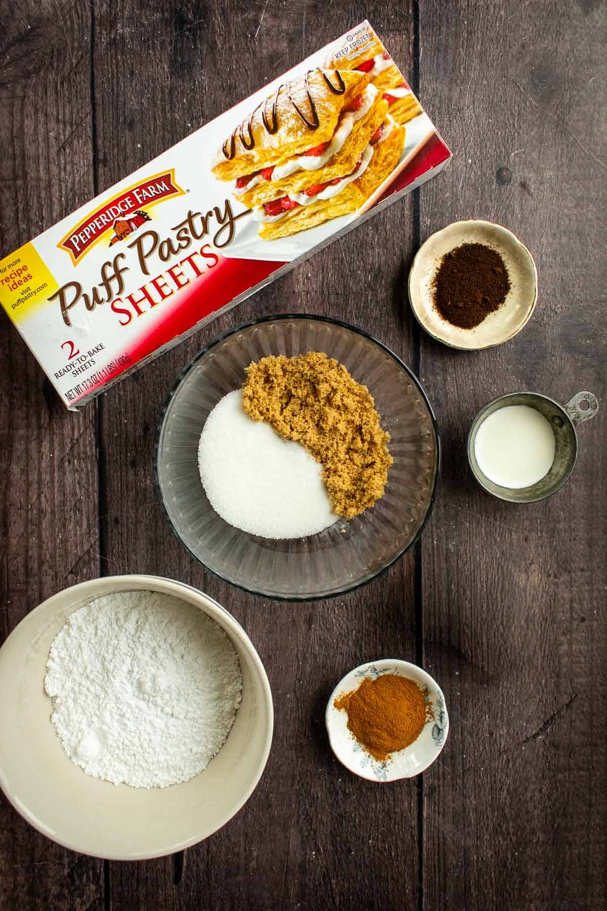 Bowls of powdered sugar, brown sugar, white sugar, espresso powder, milk, and cinnamon next to puff pastry. 