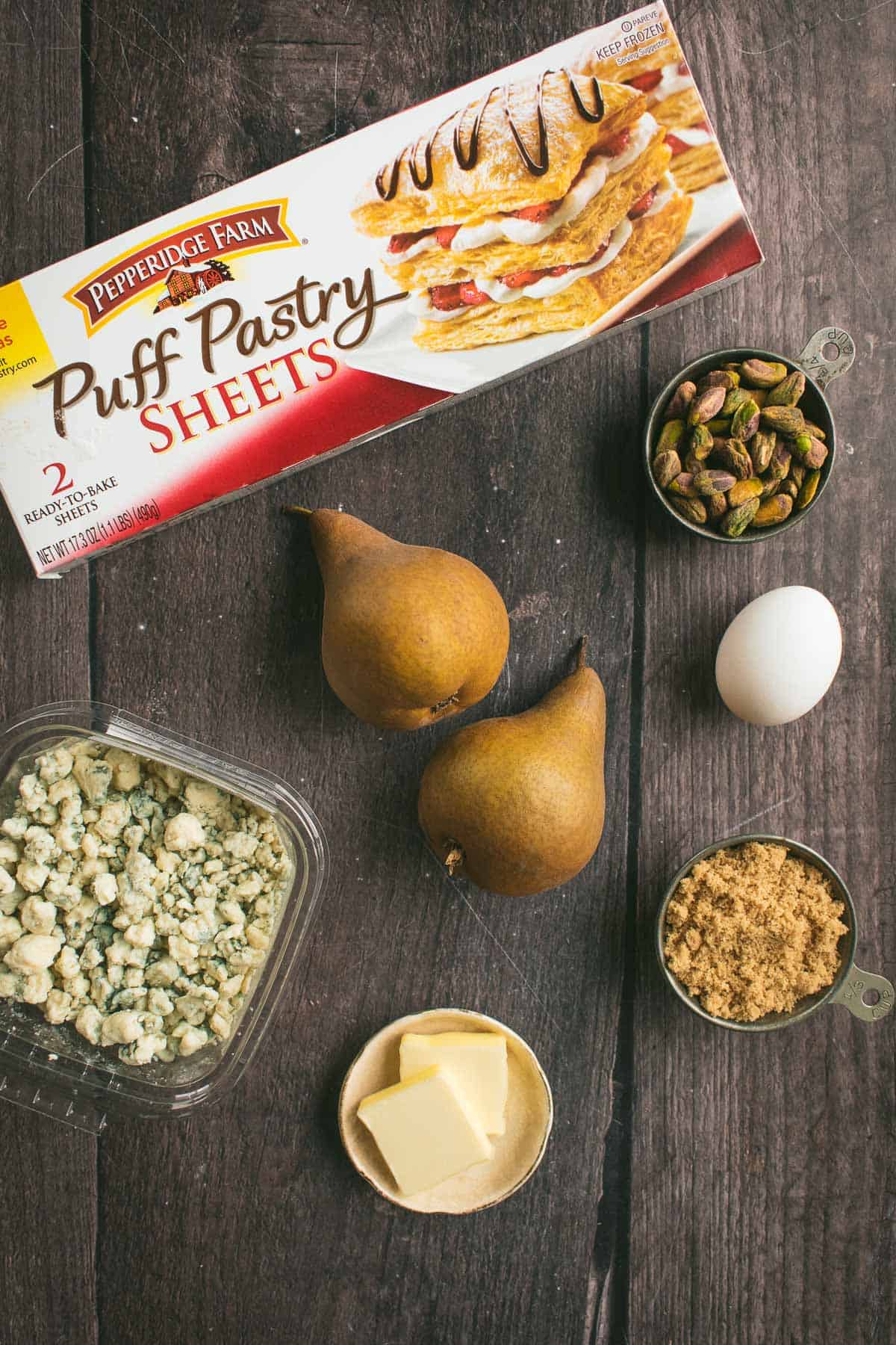 Pears on a table next to a box of puff pastry and dishes of blue cheese, pistachios, butter, brown sugar, and an egg.