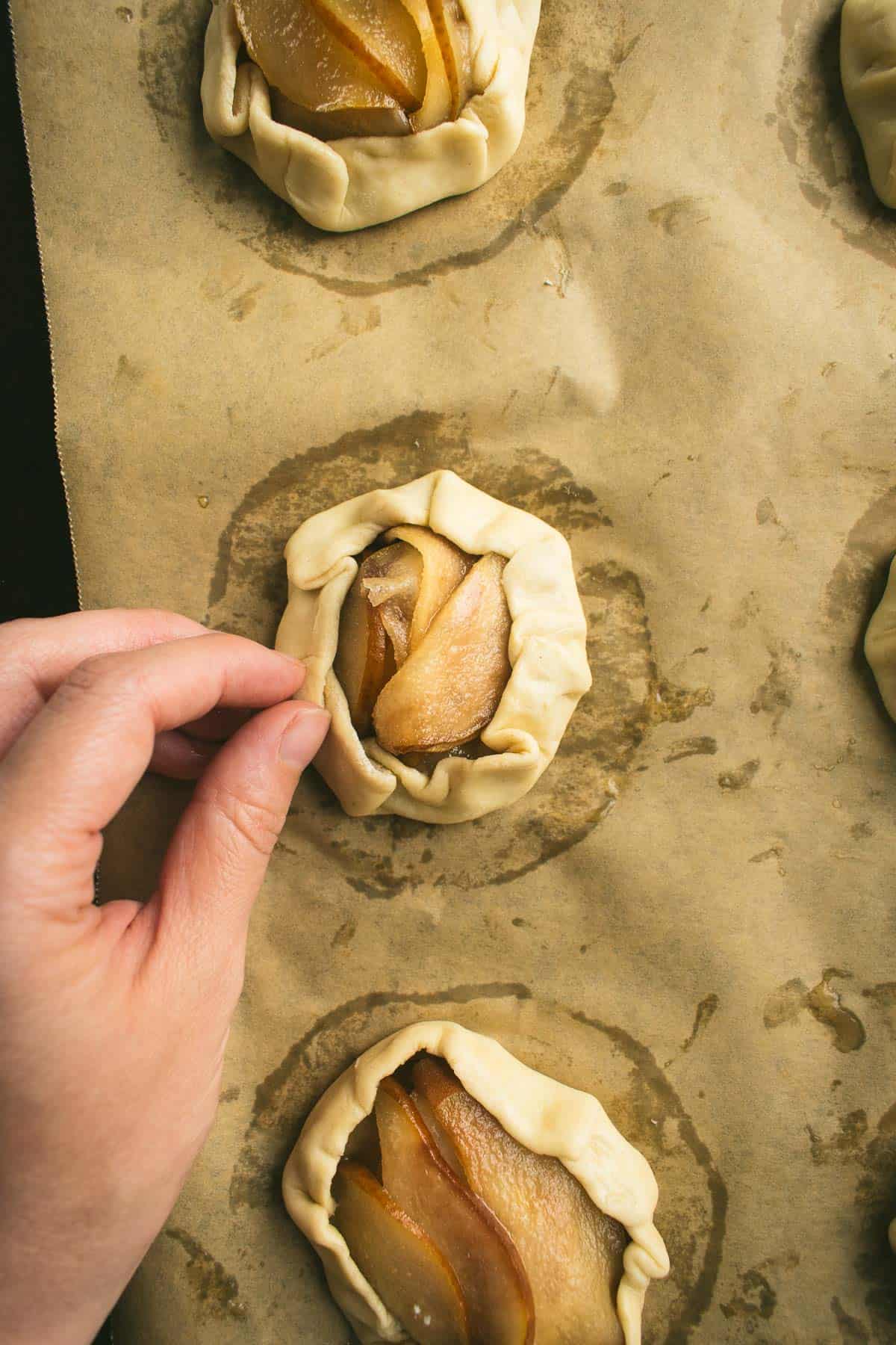 A hand folding pastry dough over pears to form mini galettes.