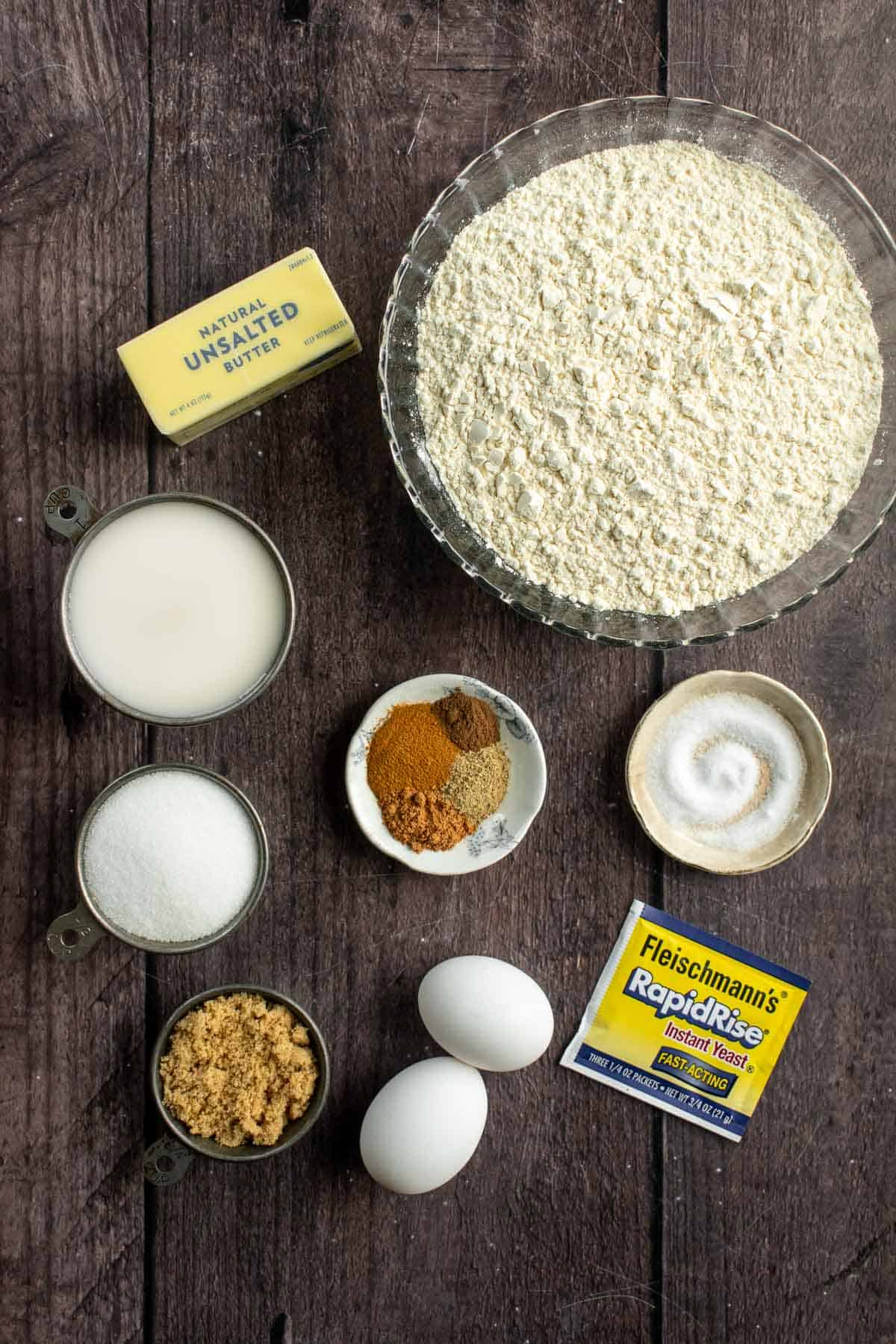 Bowls of flour, salt, spices, and sugar next to butter, eggs, and yeast on a table. 