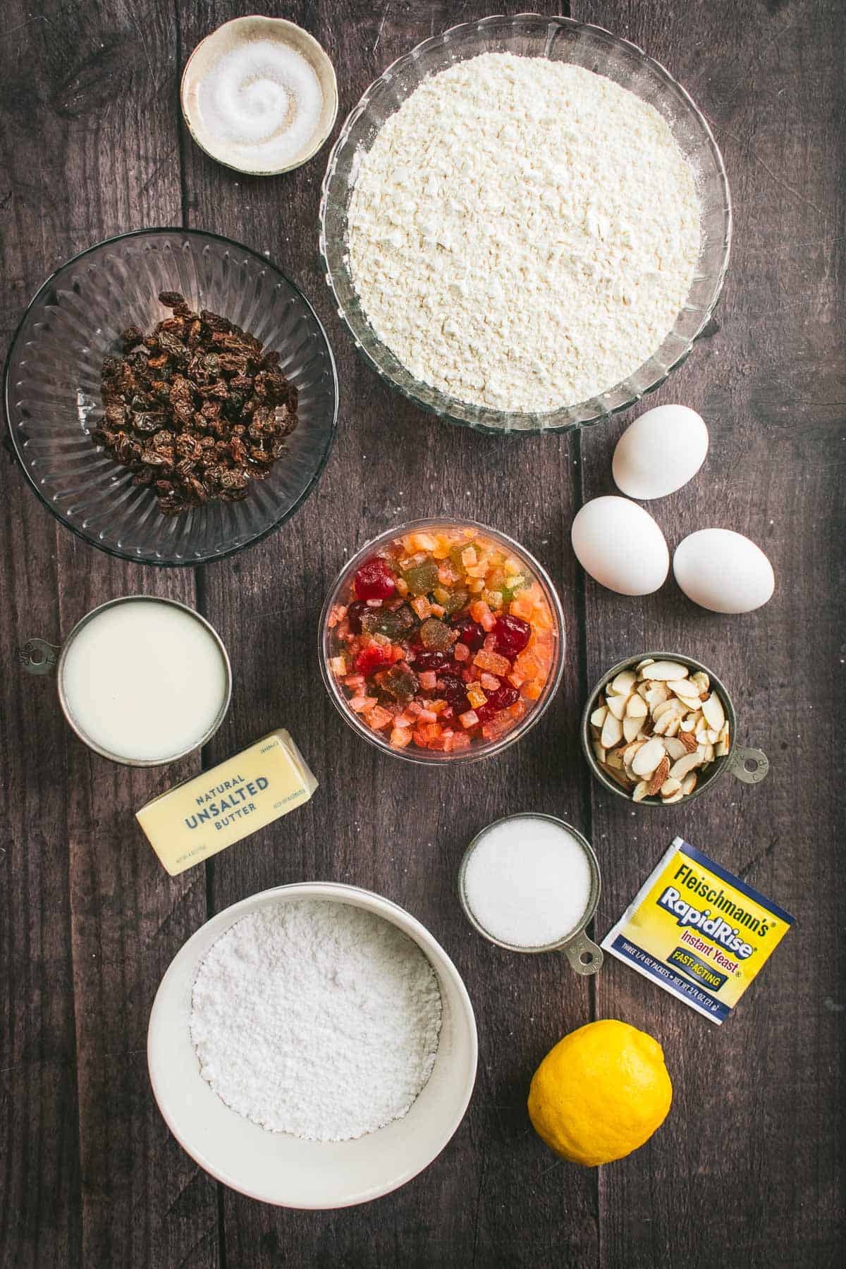 Bowls of flour, sugar, candied citron, sliced almonds, raisins, and milk next to eggs, butter, a lemon, and a packet of yeast.