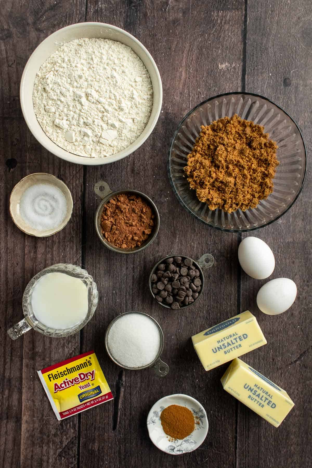 Bowls of brown sugar, flour, milk, white sugar, cinnamon, cocoa powder, and chocolate chips on a table.