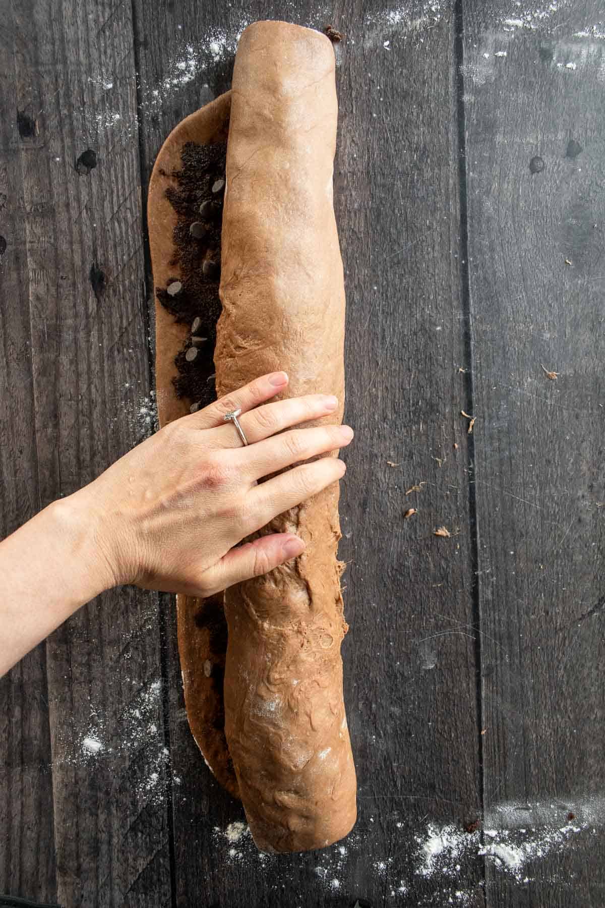 A hand rolling up a long log of brown dough on a table.