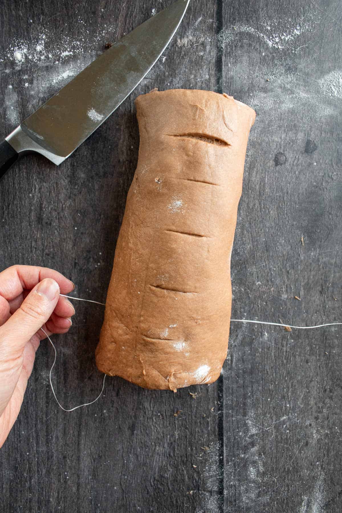 A hand cutting a log of cinnamon roll dough into rolls with dental floss.