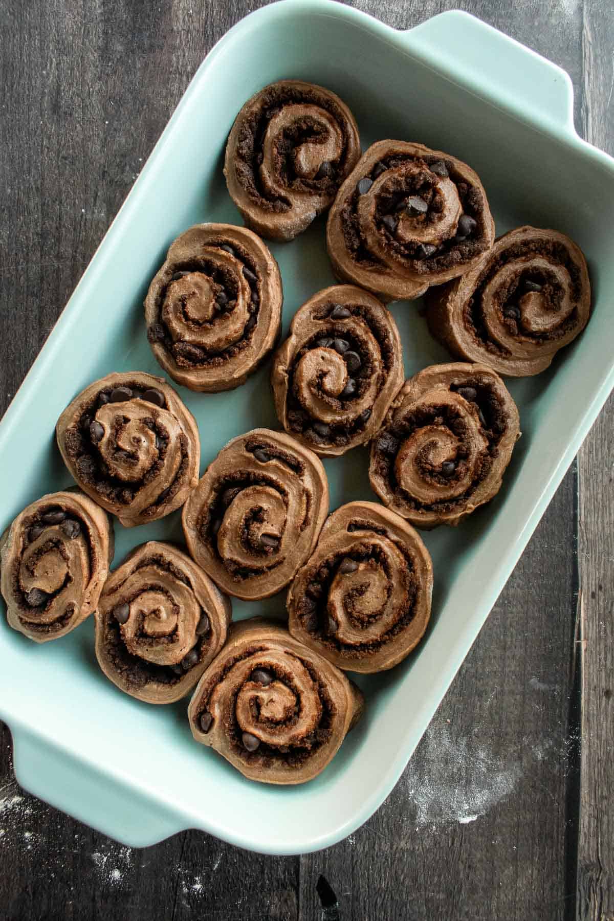 Twelve unbaked chocolate cinnamon rolls in a blue baking dish.