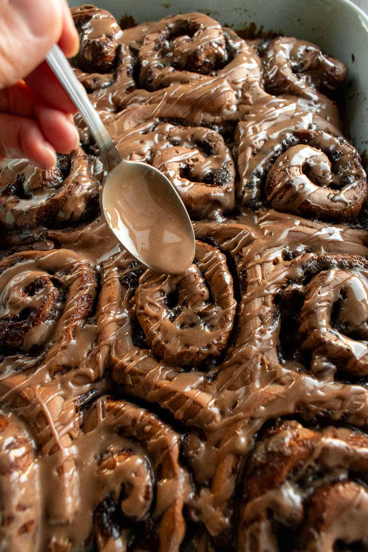A hand drizzling brown glaze from a spoon onto chocolate cinnamon buns.