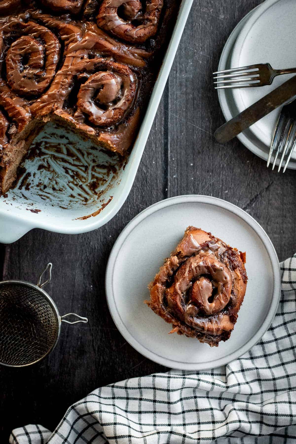 A chocolate cinnamon roll on a small plate next to more cinnamon rolls in a baking dish.