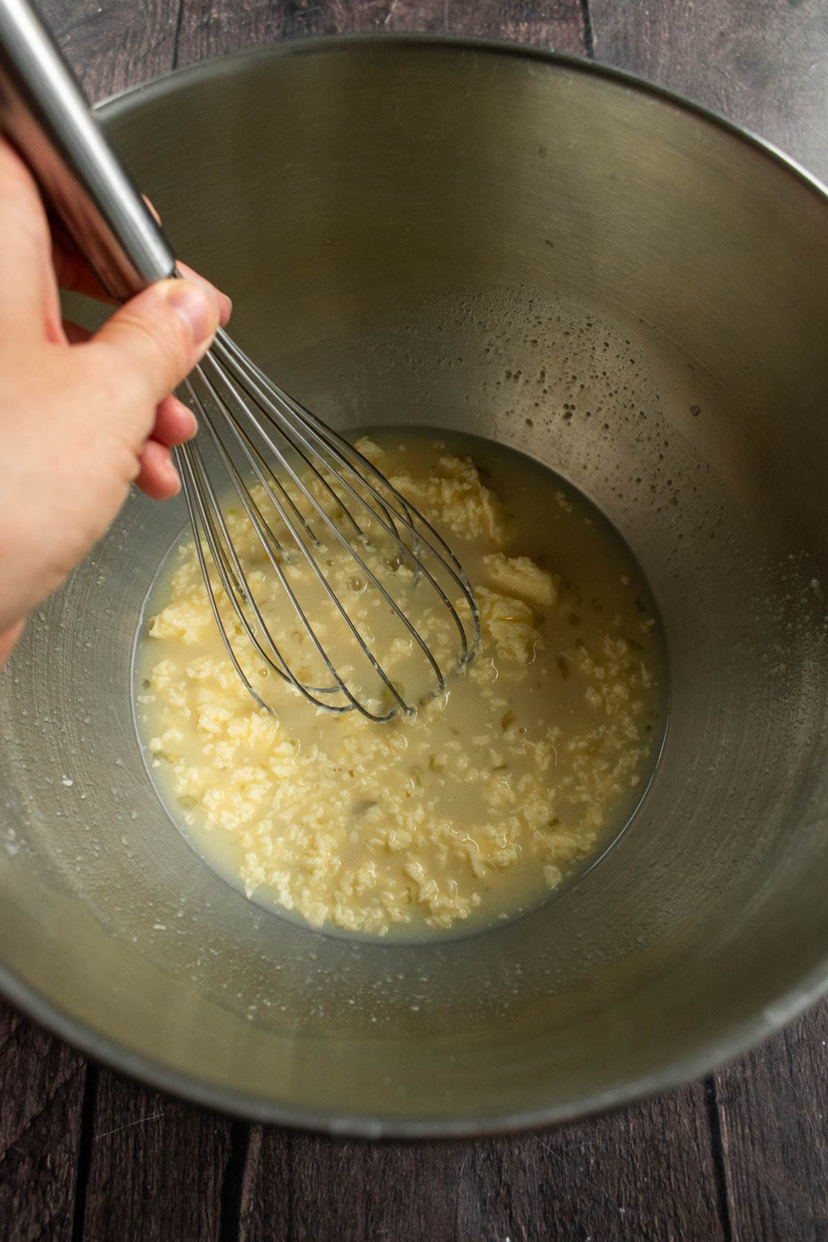 A hand whisking milk, yeast, butter, sugar, and eggs in a metal bowl.