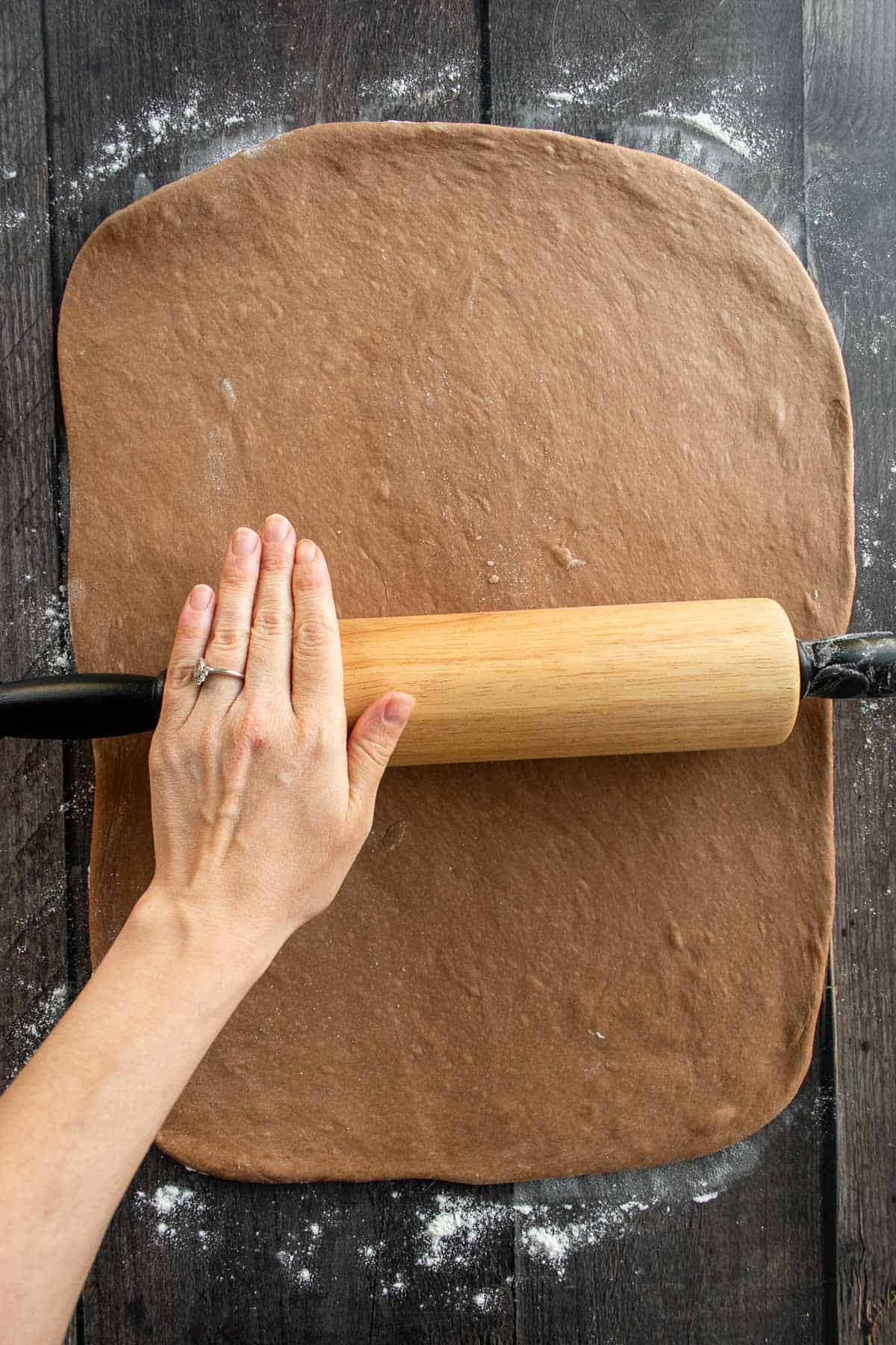 A hand rolling out brown chocolate bread dough with a rolling pin.