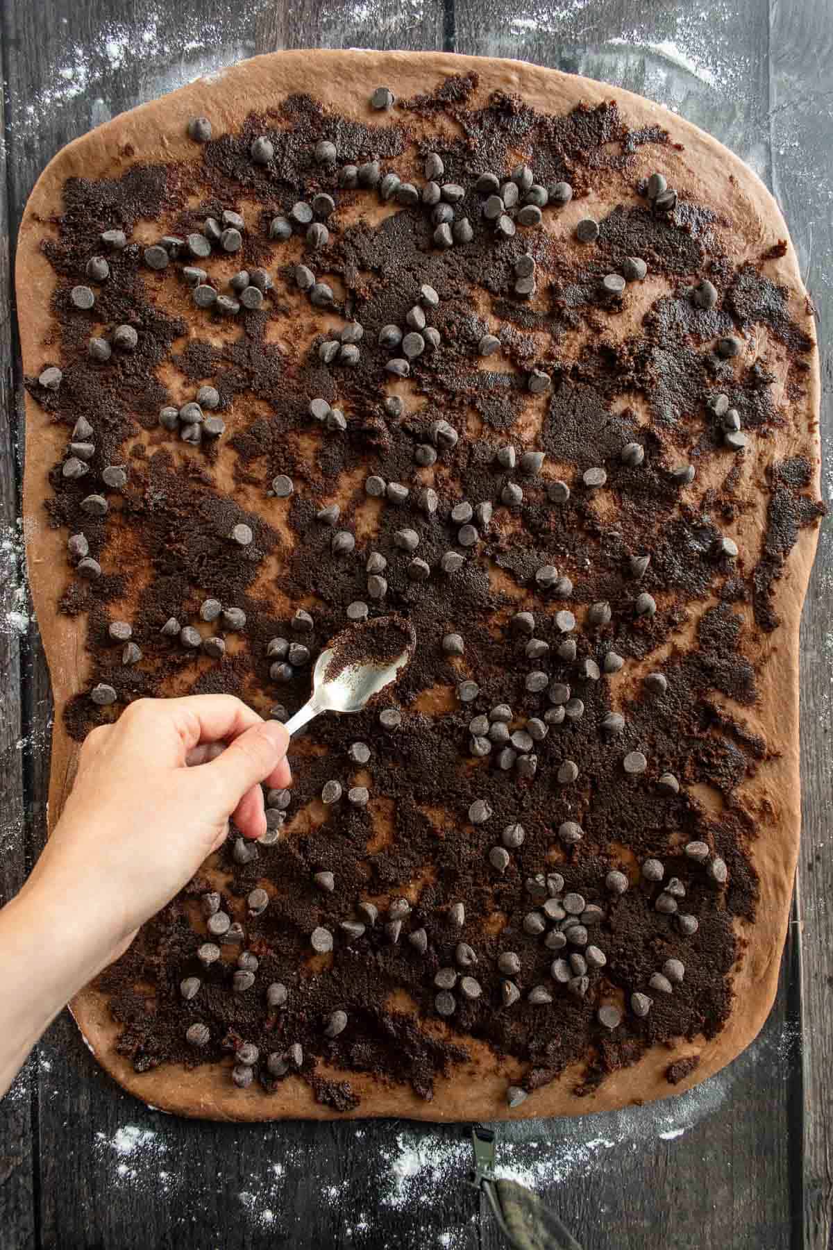 A hand spreading chocolate chips and a brown sugar filling onto chocolate cinnamon roll dough.