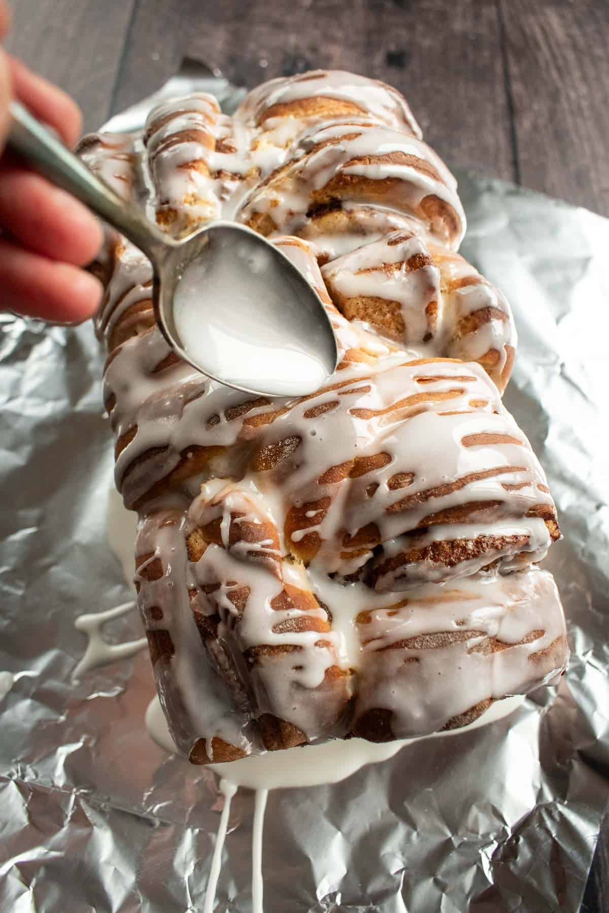 A hand drizzling white glaze from a spoon onto a baked loaf of cinnamon roll bread.