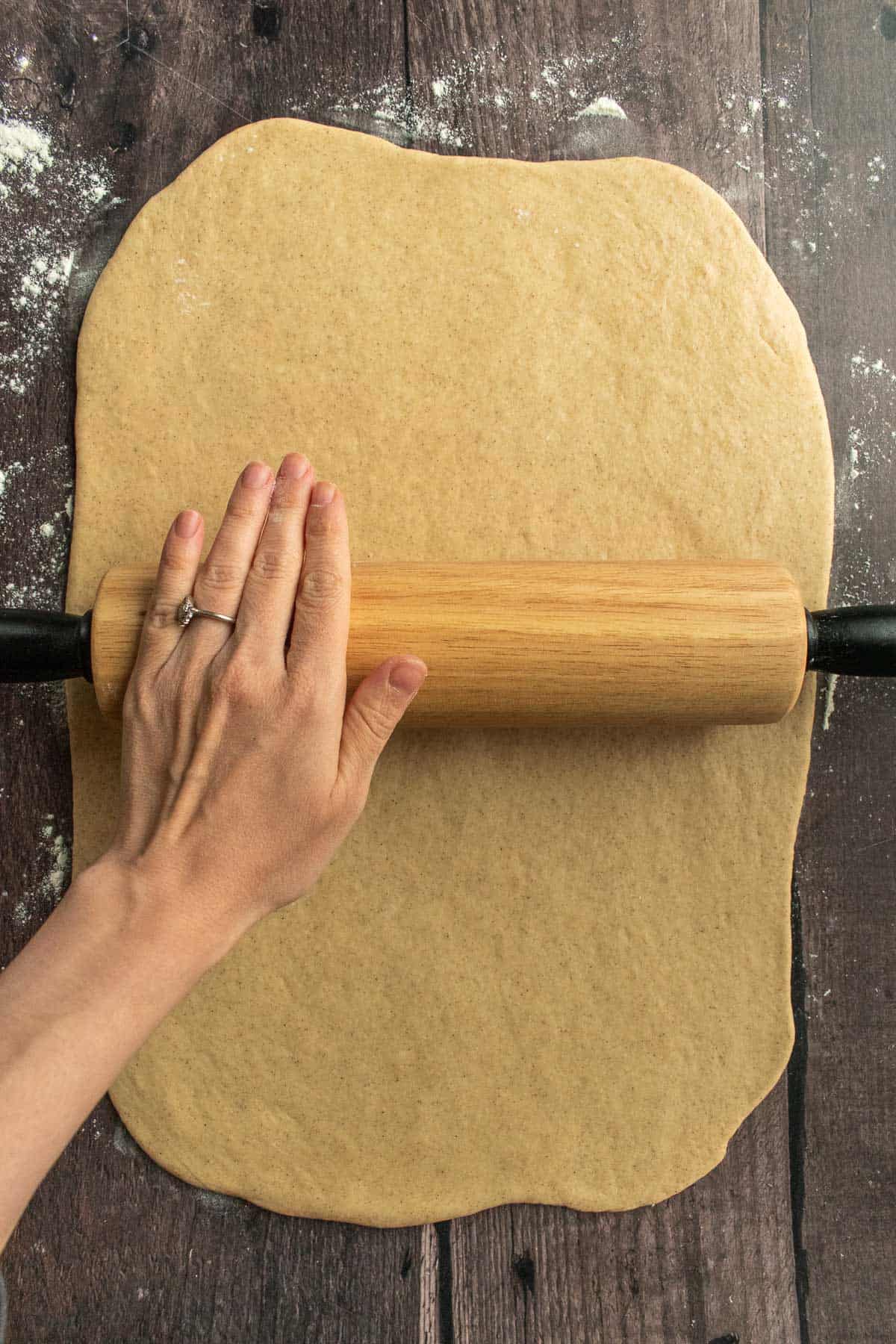 A hand using a wooden rolling pin on a large rectangle of dough.