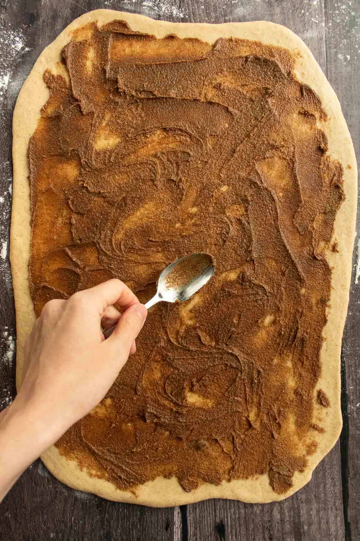 A hand spreading a creamy brown sugar mixture onto a large rectangle of dough.
