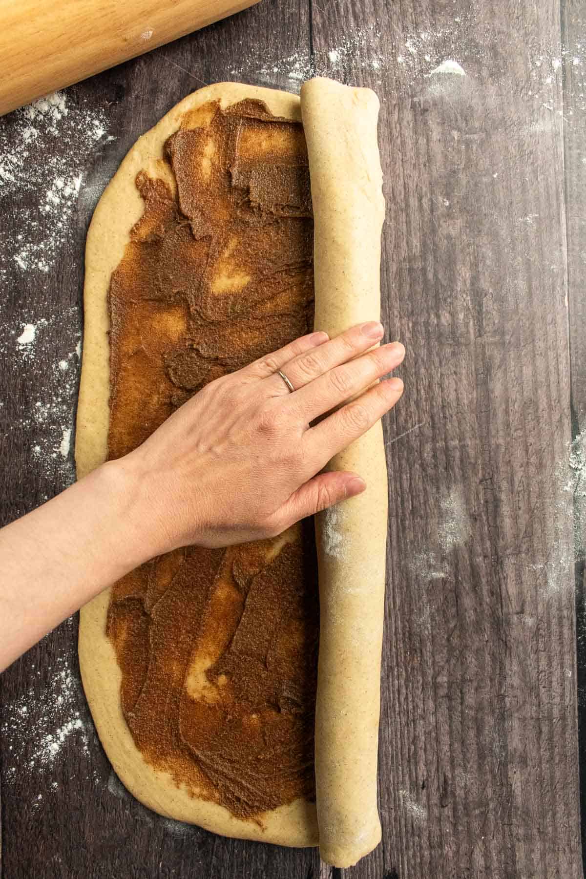 A hand rolling up bread dough filled with brown sugar and butter into a giant log.