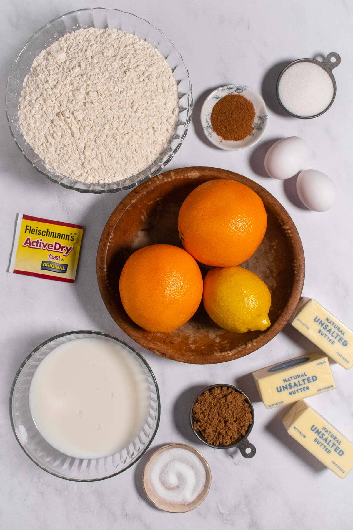 Bowls of oranges and a lemon, flour, sugar, milk, salt, and cinnamon next to eggs, butter sticks, and a packet of yeast.