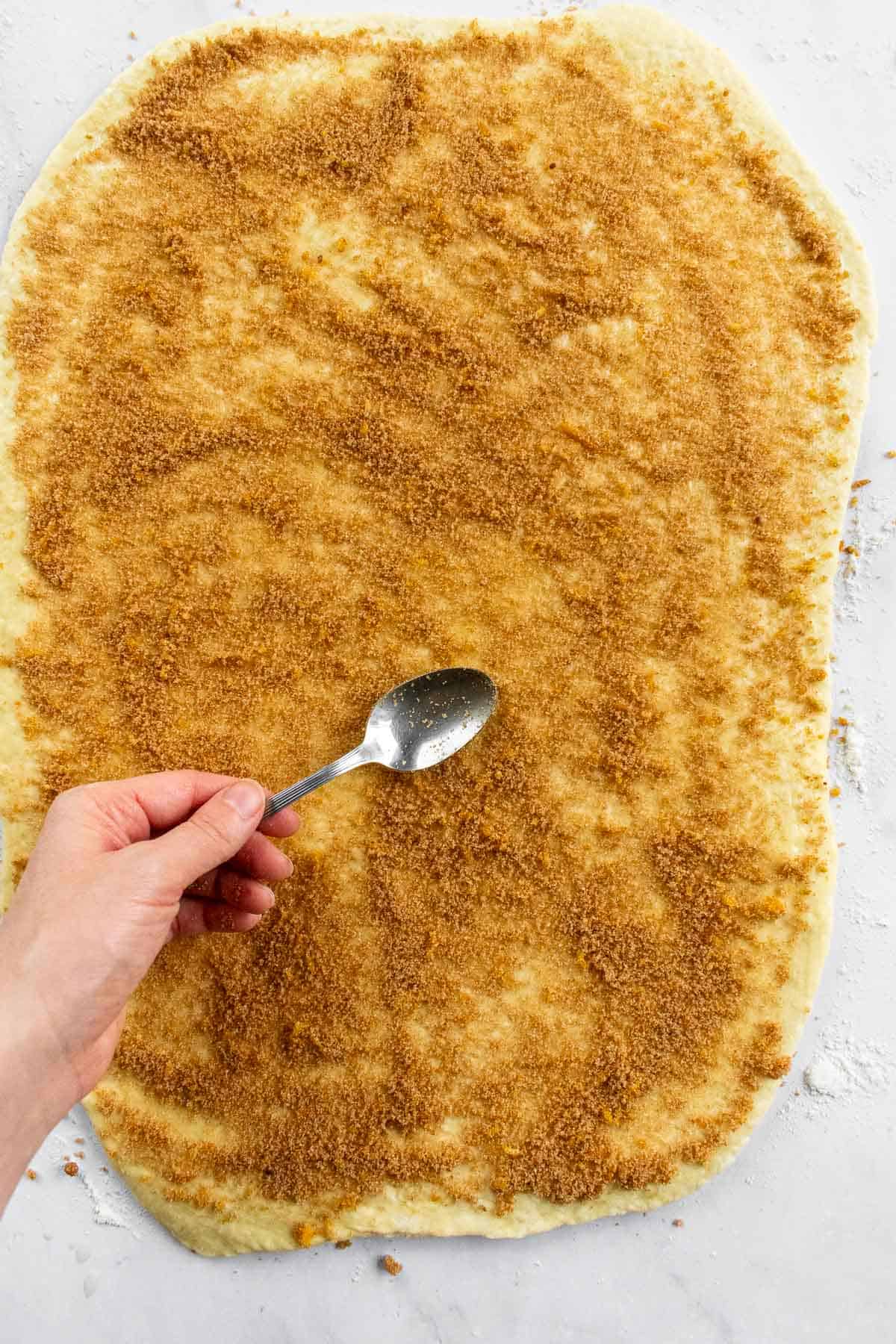 A hand spreading cinnamon sugar onto rolled-out bread dough with the back of spoon.