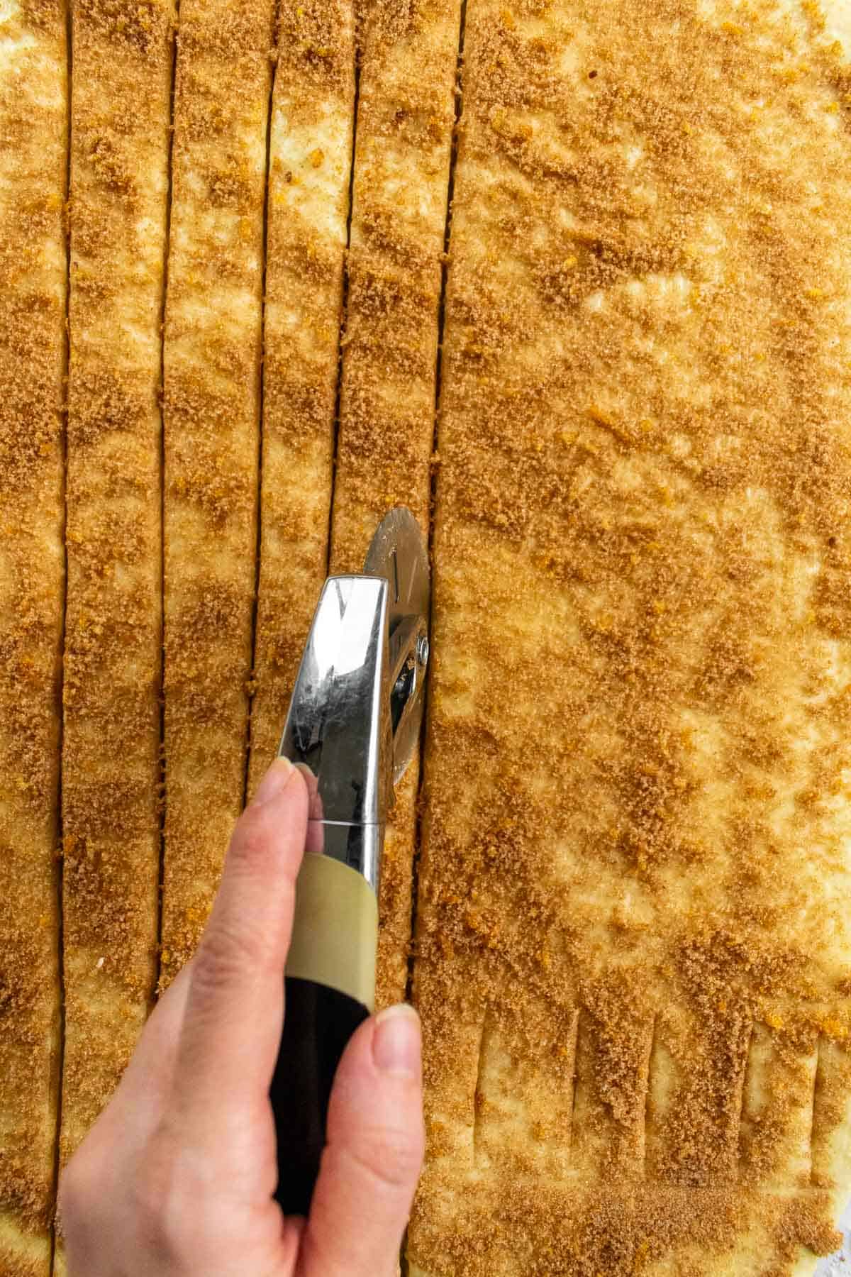 A hand using a pizza cutter to cut cinnamon-sugar-coated dough into 1-inch strips.