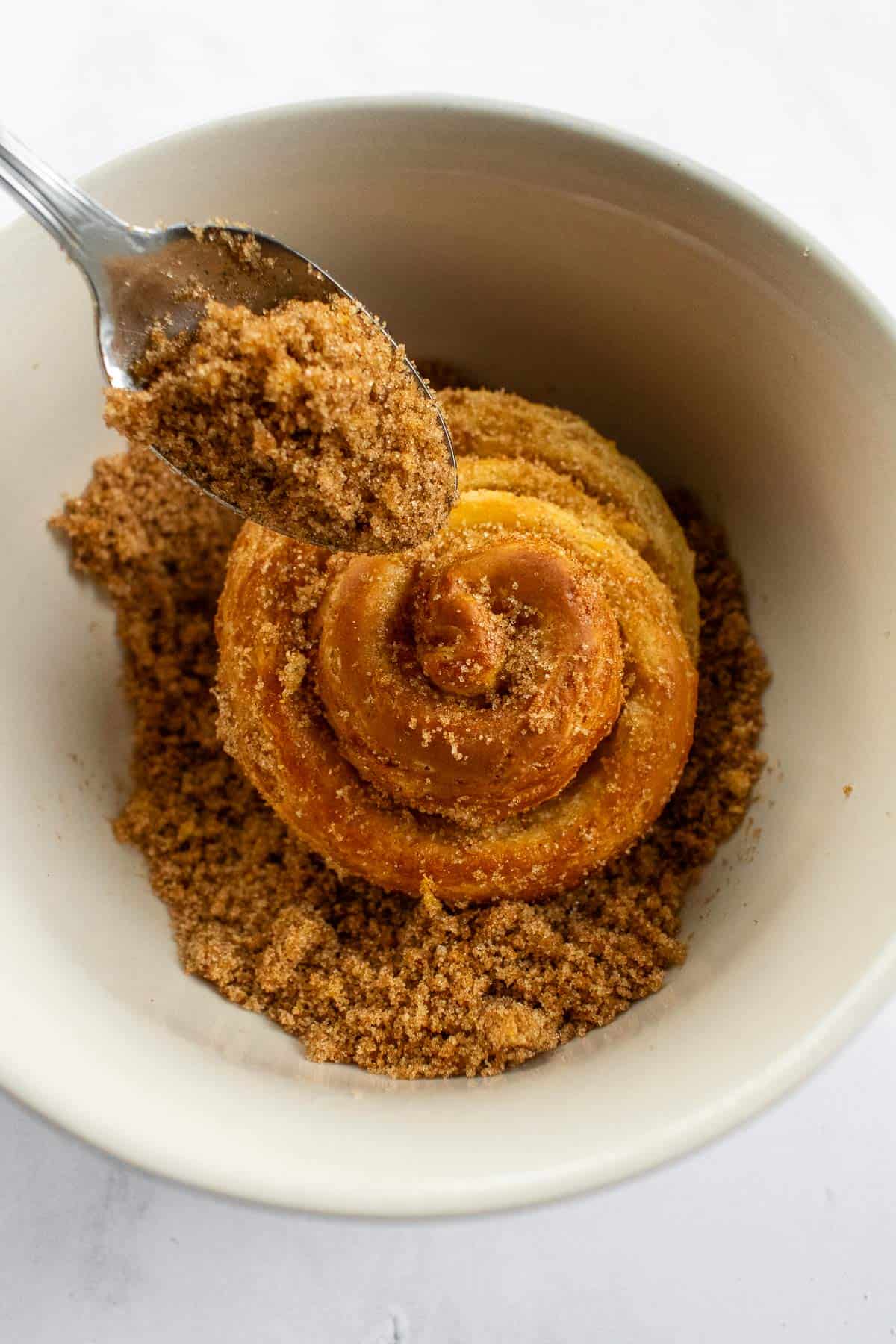 A spoon sprinkling orange-infused sugar onto a baked morning bun in a bowl.