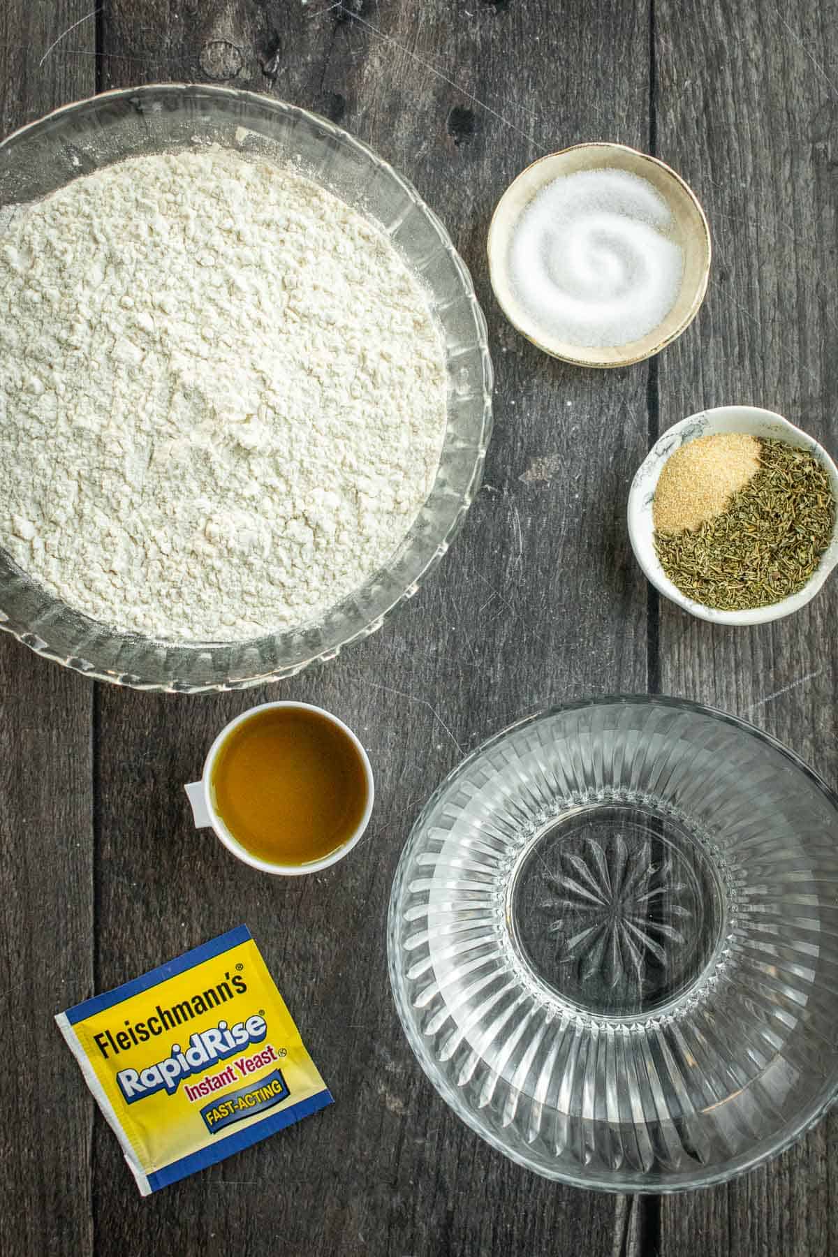 Bowls of flour, water, salt, olive oil, and herbs next to a packet of dry yeast.