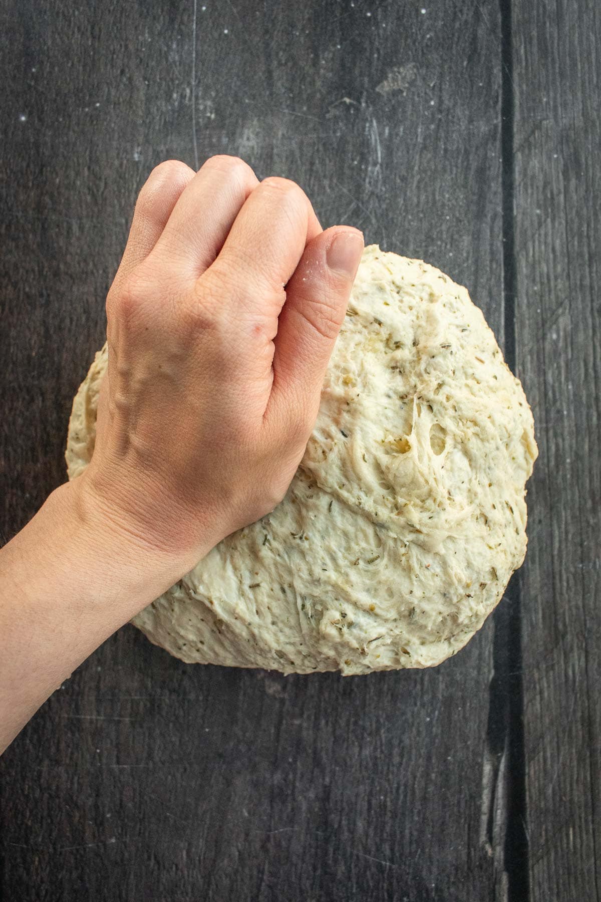 A hand kneading bread dough full of dried herbs on a table.